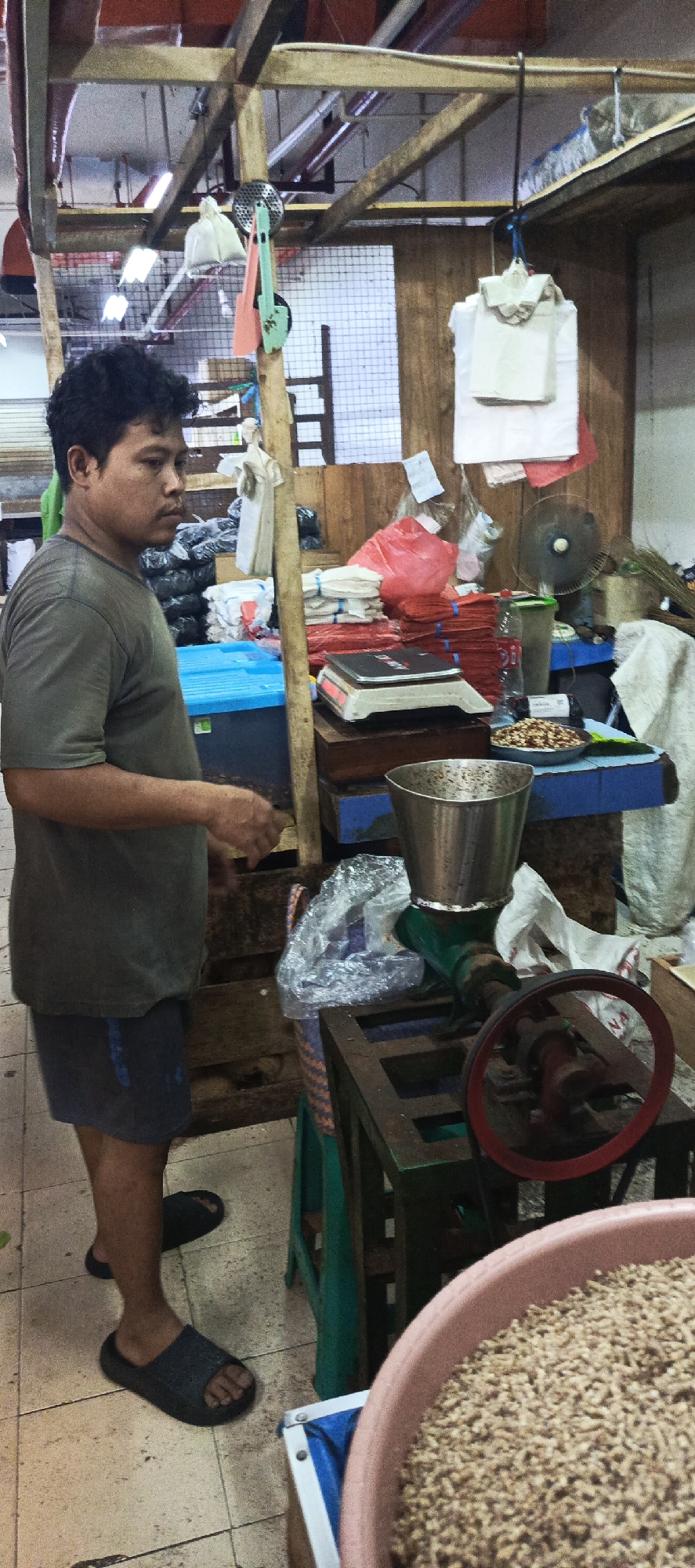 A man stands in what appears to be a market stall in Sukaresmi, Indonesia. He is wearing a grey t-shirt and dark shorts, and dark sandals. He is looking to his left, his right hand slightly raised and extended. Behind him, a wooden stall structure is visible, with various items for sale and display. To the man's left, there are stacks of black and white plastic bags. In the center of the stall, there are piles of red plastic bags and white bags, as well as a scale and a blue plastic tub. To the right of the man, a small green machine with a metal funnel and a red wheel is present. A large pink basin filled with small, light-brown pellets is in the foreground, partially obscuring the machine. The stall appears to be a retail environment, likely selling goods such as bags or food products, given the presence of the pellets and scale.
