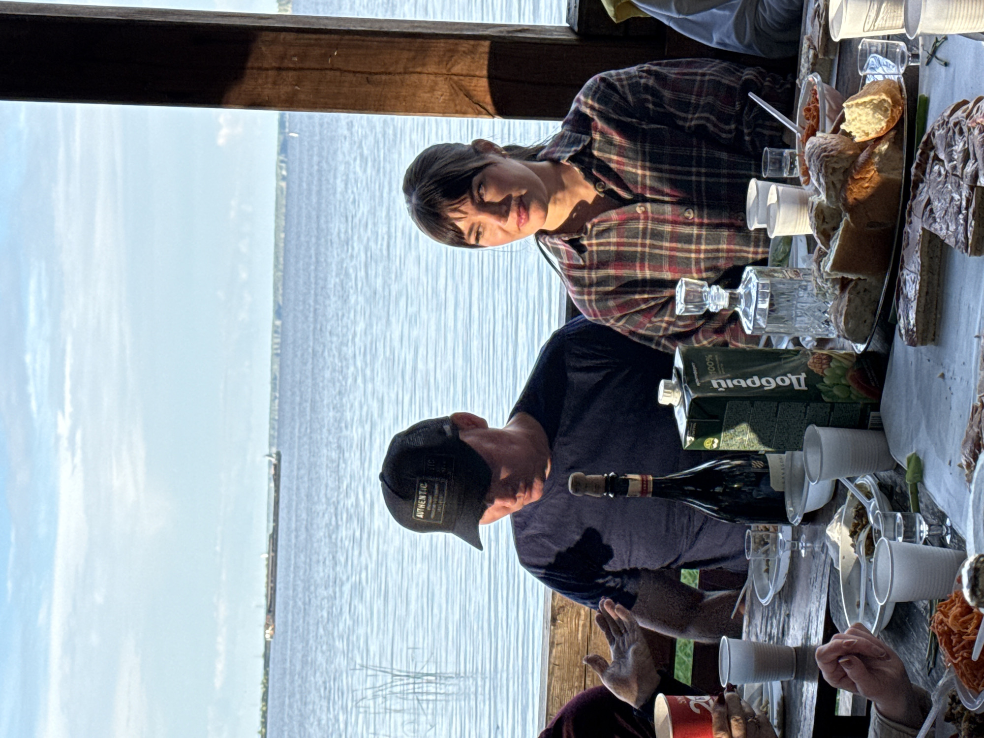 A man and a woman are seated at a wooden table outdoors by a body of water. The man, wearing a dark blue t-shirt and a black baseball cap, is looking down. The woman, in a plaid shirt, is looking to her right. On the table are plates of food, bread, a bottle of wine, a carton of juice, glasses, and plastic cups. In the background, the water stretches to the horizon under a partly cloudy sky. A wooden structure is visible on the right side of the image.