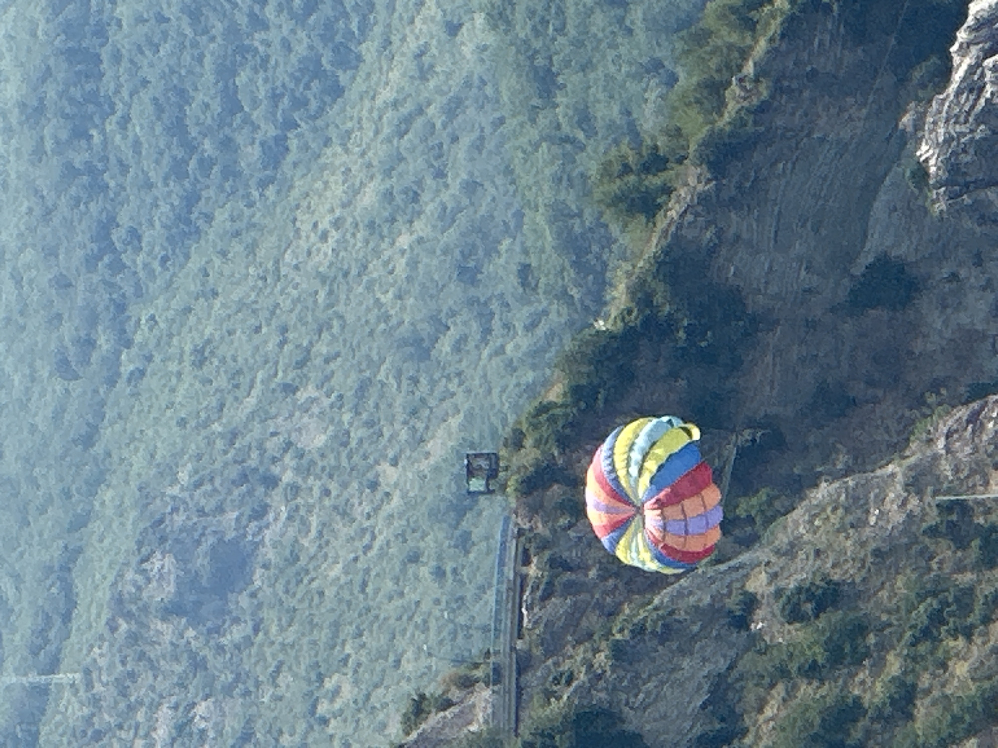 This is a high-angle, vertical shot of a colorful paraglider suspended in the air against a backdrop of lush, green, mountainous terrain. The paraglider, with its vibrant sections of red, orange, yellow, blue, and purple, is the central object of focus, appearing to be in mid-descent or ascent. Below the paraglider, a steep, rocky cliff face is visible, dotted with vegetation. Further down the mountainside, there is a glimpse of a road or path and a small, dark rectangular structure, possibly a building or lookout point. The overall scene suggests an outdoor recreational activity in a natural, mountainous setting, likely in Boreti, Montenegro. The lighting indicates it is daytime, with clear visibility. The image evokes a sense of adventure and freedom associated with paragliding. There is no visible text in the image.