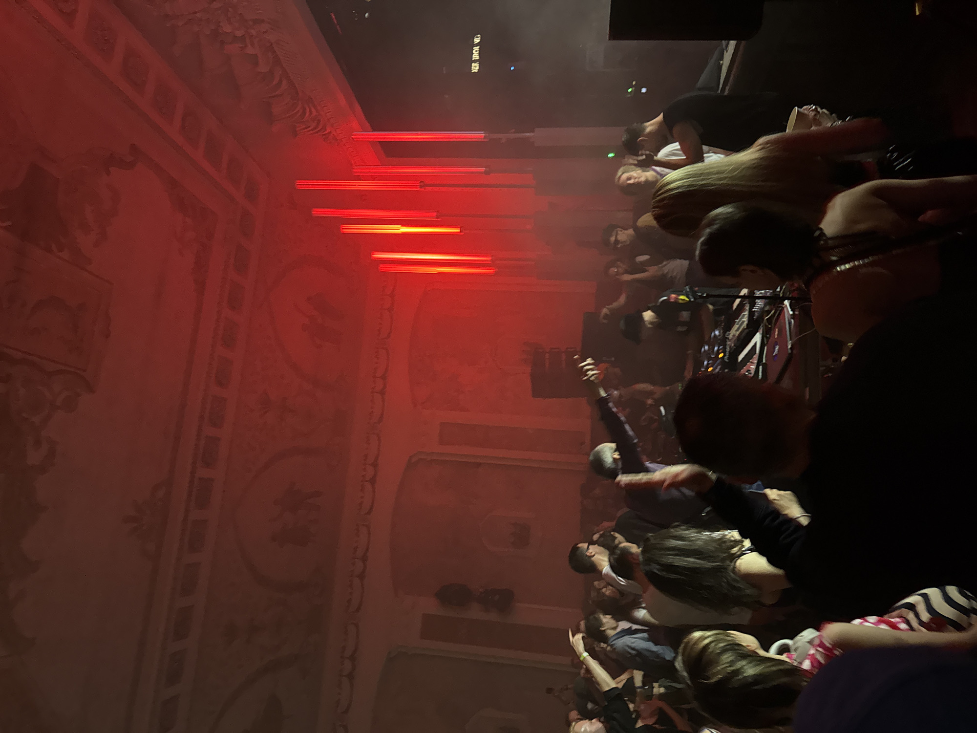 The image captures an active indoor event, likely a music performance or club night, held within an ornate historical building in Khimki, Russia. The scene is dominated by a pervasive red light that bathes the entire space.
The venue features high ceilings and walls adorned with classical architectural elements, including decorative moldings, arches, and faint frescoes depicting figures and patterns, visible despite the dim, monochromatic lighting. Suspended from the ceiling or positioned vertically as part of the stage setup are several tall, cylindrical red light tubes, forming a prominent visual display. Haze or smoke is present, illuminated by the red light, adding to the atmospheric effect. On the far right, a large dark screen or wall displays partially legible white text, possibly "SER IRON UG".
A large crowd of people, mostly adults, occupies the foreground and mid-ground, facing towards a central area where a DJ setup is located. Individuals are standing, some with arms raised, indicating engagement with the event. In the middle left, a person is visible with their right arm extended and pointing. The DJ station, situated in the mid-ground, features turntables, a mixer, and what appears to be a laptop or control interface, with a person actively engaging with the equipment. People in the foreground are largely seen from behind, including one individual wearing a horizontally striped top. Visible interactions primarily involve standing, watching, and general movement within the crowd.