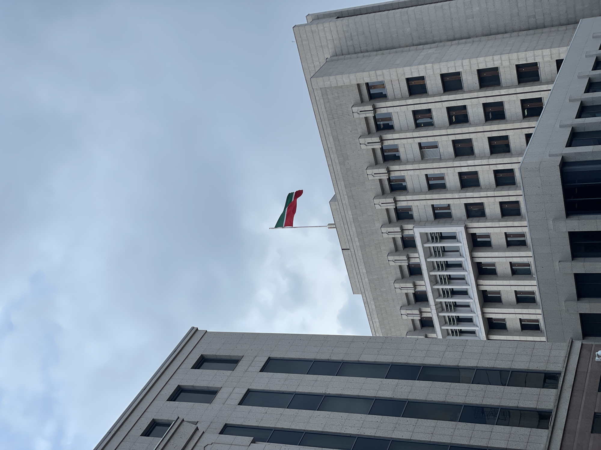 An upward-angled photograph captures the angular facades of two modern, grey stone-clad buildings against an overcast sky. A flag with vertical green, white, and red stripes, likely the national flag of Mexico or Italy, flies from a pole in the space between the structures.
The building on the right dominates the upper half of the frame, showcasing a stepped or terraced design with multiple stories of square, recessed windows. Its concrete or stone exterior features repetitive geometric patterns and strong horizontal and vertical lines, giving it a robust, Brutalist or post-modern appearance.
Part of a second, similar building is visible in the lower-left corner. This building also presents a grey, stone-clad facade with rectangular windows, some wider than others, arranged horizontally. Its clean lines and material selection complement the larger structure.
The sky is uniformly grey and cloudy, suggesting an overcast day with soft, diffuse light. No people or discernible text are visible in the image. The perspective emphasizes the towering presence and architectural details of the urban environment.