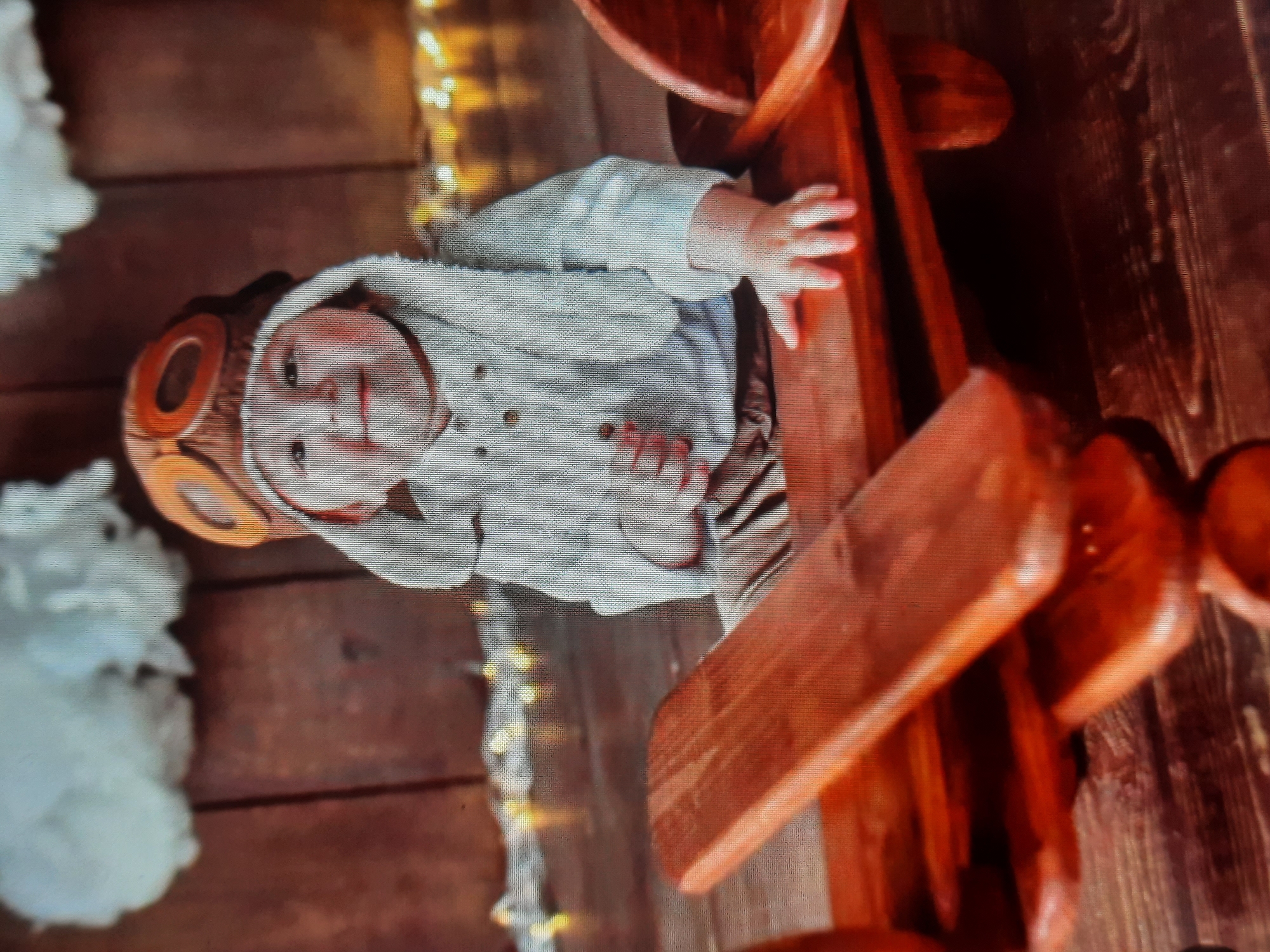 A young child, appearing to be a toddler, is seated on a wooden airplane prop. The child is wearing an aviator hat with goggles and a light-colored shirt with buttons. The background consists of a wooden wall with fluffy white decorations at the top and a string of yellow lights along the bottom. The floor is also made of dark wood planks. The image is a vertical, slightly blurry photograph, likely taken indoors in a studio setting. There are no identifiable landmarks or context to suggest a specific location in Rosenheim, Germany.