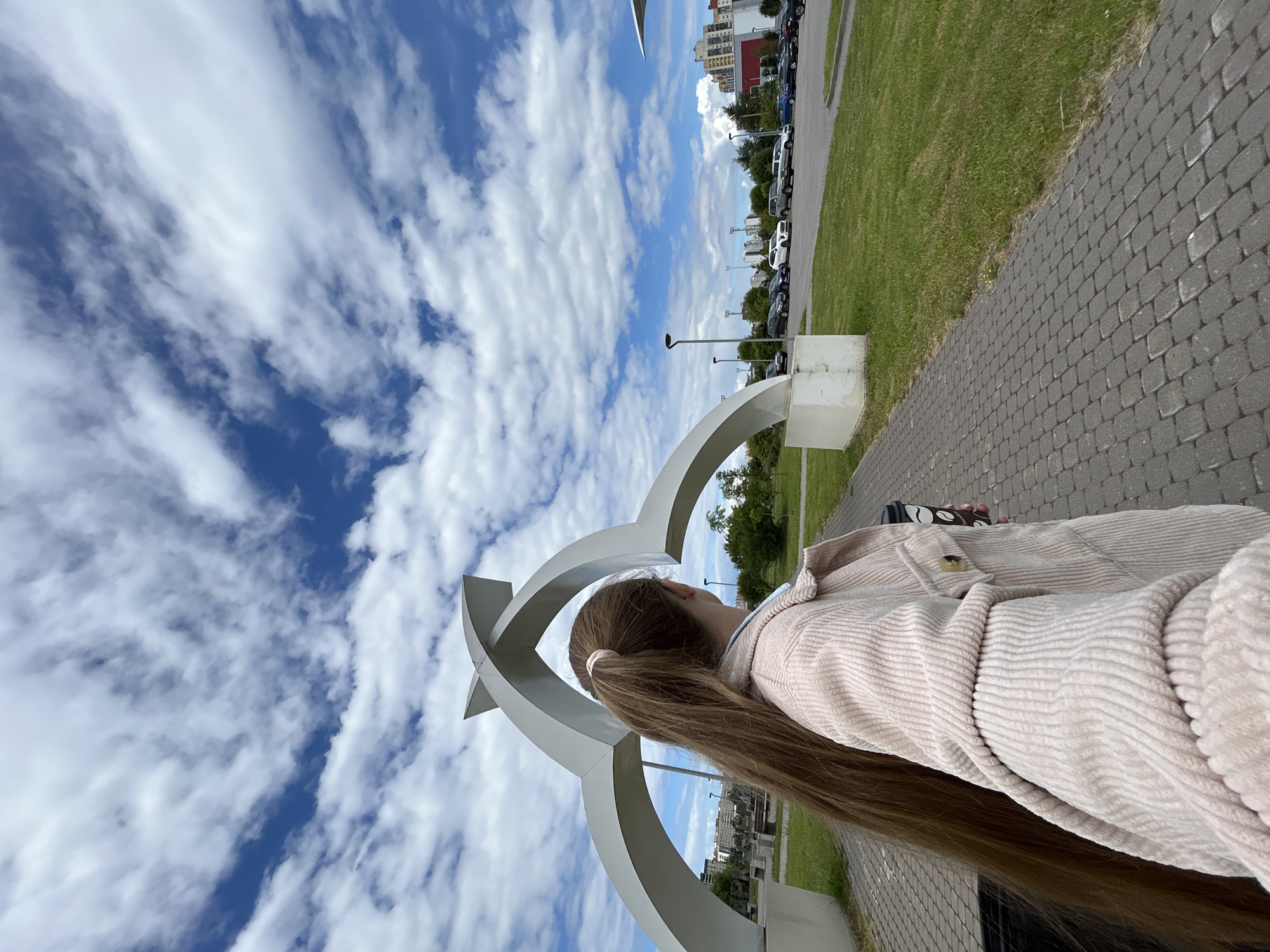A young woman with long, light brown hair tied in a ponytail, facing away from the camera, stands on a paved walkway in front of a large, white, abstract sculpture. She is wearing a light pink corduroy jacket and holding a disposable coffee cup. The sculpture, which appears to be made of three interlocking arches, is situated in an outdoor public space. Beyond the sculpture and walkway, there is a well-maintained lawn, a parking lot filled with various cars, streetlights, and in the background, buildings suggesting an urban environment with a clear blue sky dotted with white, wispy clouds. The scene is likely a public park or plaza within the city of Saint Petersburg, Russia.