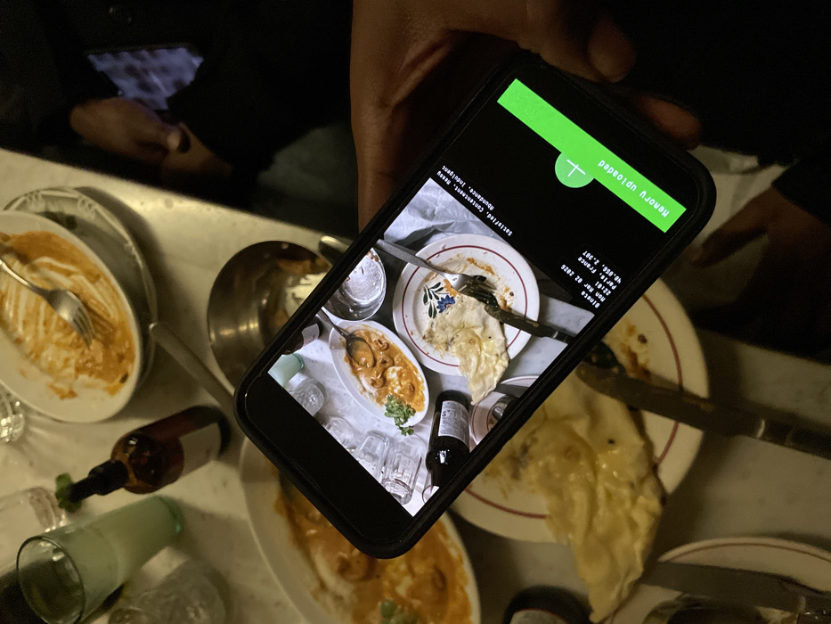 A person's right hand holds a black smartphone, tilted diagonally, dominating the central foreground of the image. The smartphone screen displays an overhead photograph of a dining table with partially eaten food, specifically a white oval dish containing a red-orange curry with shrimp-like contents, a white plate with a piece of naan bread and a fork, and an empty glass. Text overlays on the smartphone screen indicate "Binsta", "Mon Mar 02 2020", "22:01", "Paris, France", and coordinates "48.856, 2.351". Additional text reads "Satisfied, Contentment, Peony Abundance, Indulgent" and a green bar labeled "Memory uploaded".
The physical scene in the background depicts a marble-like dining table in a dimly lit restaurant setting. Visible items on the table include a white oval dish with an orange curry, a partially eaten plate of naan bread, a mostly empty oval dish with residual orange sauce and a fork, a tall green glass containing a light beverage, a dark brown pump bottle, and a metallic bowl. A dark-clothed arm and a hand holding another reflective phone are visible in the blurred background to the right. The location, as specified on the smartphone display, is Paris, France, with coordinates 48.856, 2.351, which corresponds to the central area of Paris near the Seine river, specifically in the vicinity of the Île de la Cité.