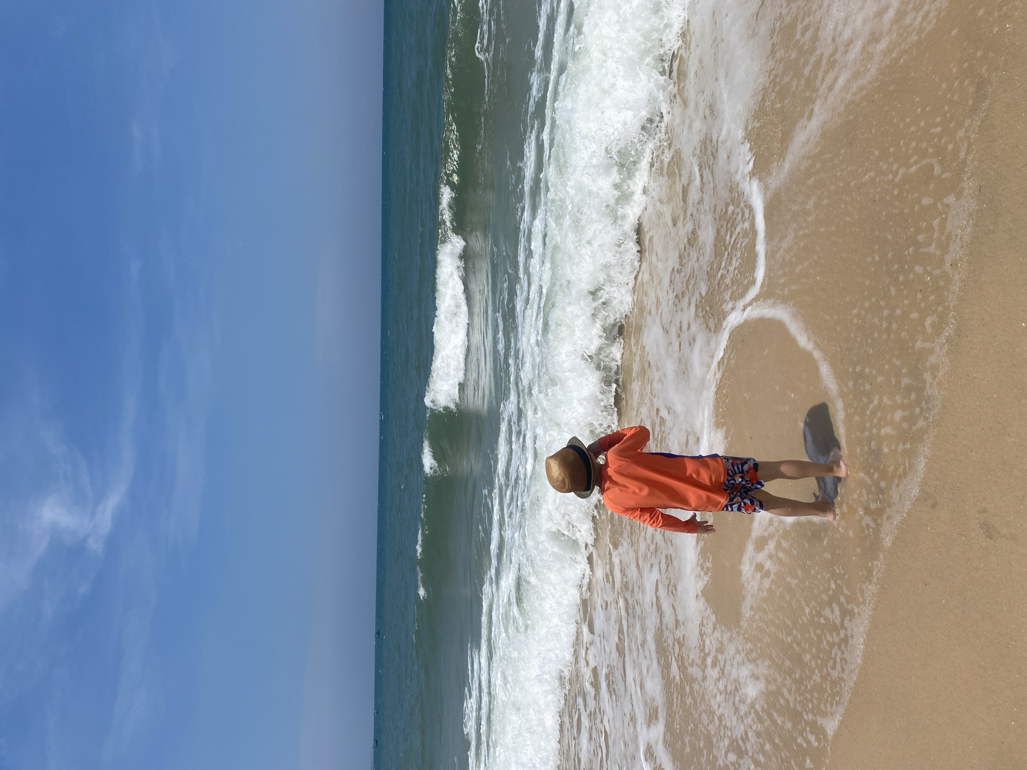 The image depicts a person standing on the beach at Phu Thuy Ward, Vietnam. The individual is wearing an orange long-sleeved shirt, patterned shorts, and a straw hat. They stand near the edge of the water, with ocean waves washing onto the sandy shore. The sky is blue and clear.