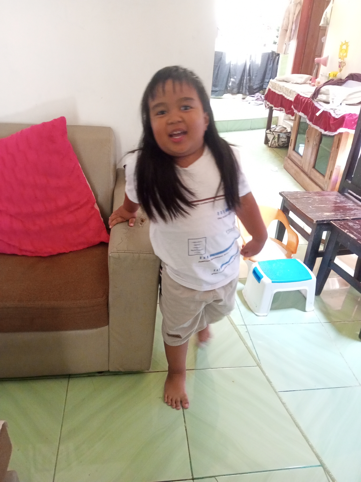 A young child with long, dark hair stands in a room in Krasak, Indonesia, with a beige and brown sofa to their left and a wooden table with two stools to their right. The child is wearing a white t-shirt with blue graphics and light beige shorts. Their mouth is open in a smile, and their right hand rests on the armrest of the sofa. In front of the child, there is a small white and blue plastic stool. The floor is tiled in a light green color. In the background, a wooden cabinet with glass doors is visible, along with a table covered with a red and white cloth. The room appears to be a domestic setting.