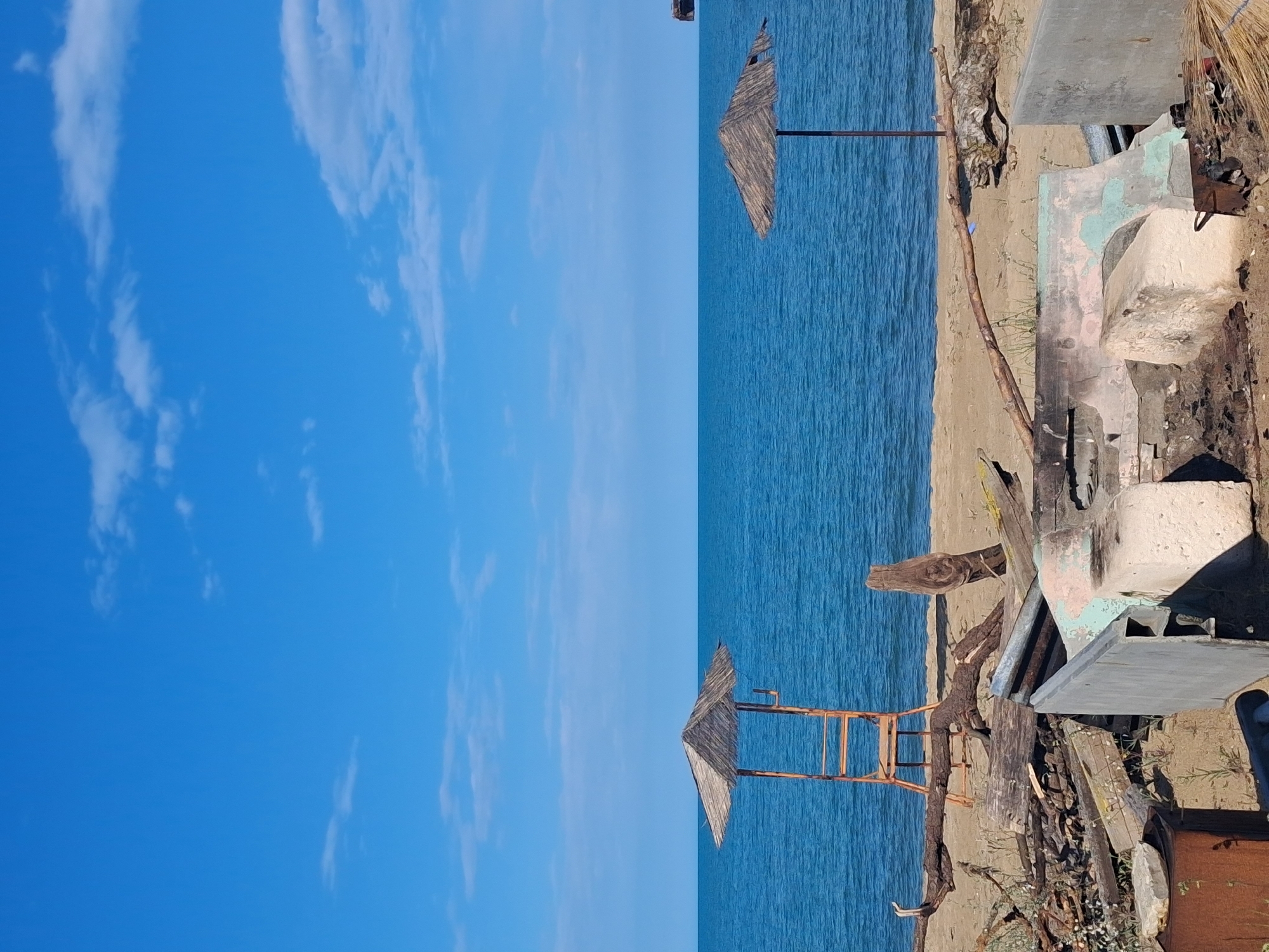 This image captures a beach scene in Shkorpilovtsi, Bulgaria. The upper third of the frame shows a clear blue sky with scattered white cumulus clouds. The middle third displays a calm, deep blue sea extending to a flat horizon. The foreground consists of a sandy beach littered with various objects. On the left, a rudimentary wooden and thatched-roof structure, resembling an observation post or lifeguard chair, stands partially on the sand. Extensive driftwood, logs, and assorted debris are scattered across the left side of the foreground, including a rusted metal container in the lower-left corner. On the right, a single thatched beach umbrella is rooted in the sand near the water's edge. A substantial, broken concrete or stone structure with residual light green paint is visible in the right foreground, surrounded by additional driftwood and debris. A dark, indeterminate object is present on the water surface in the distant right. No persons are depicted within the scene.