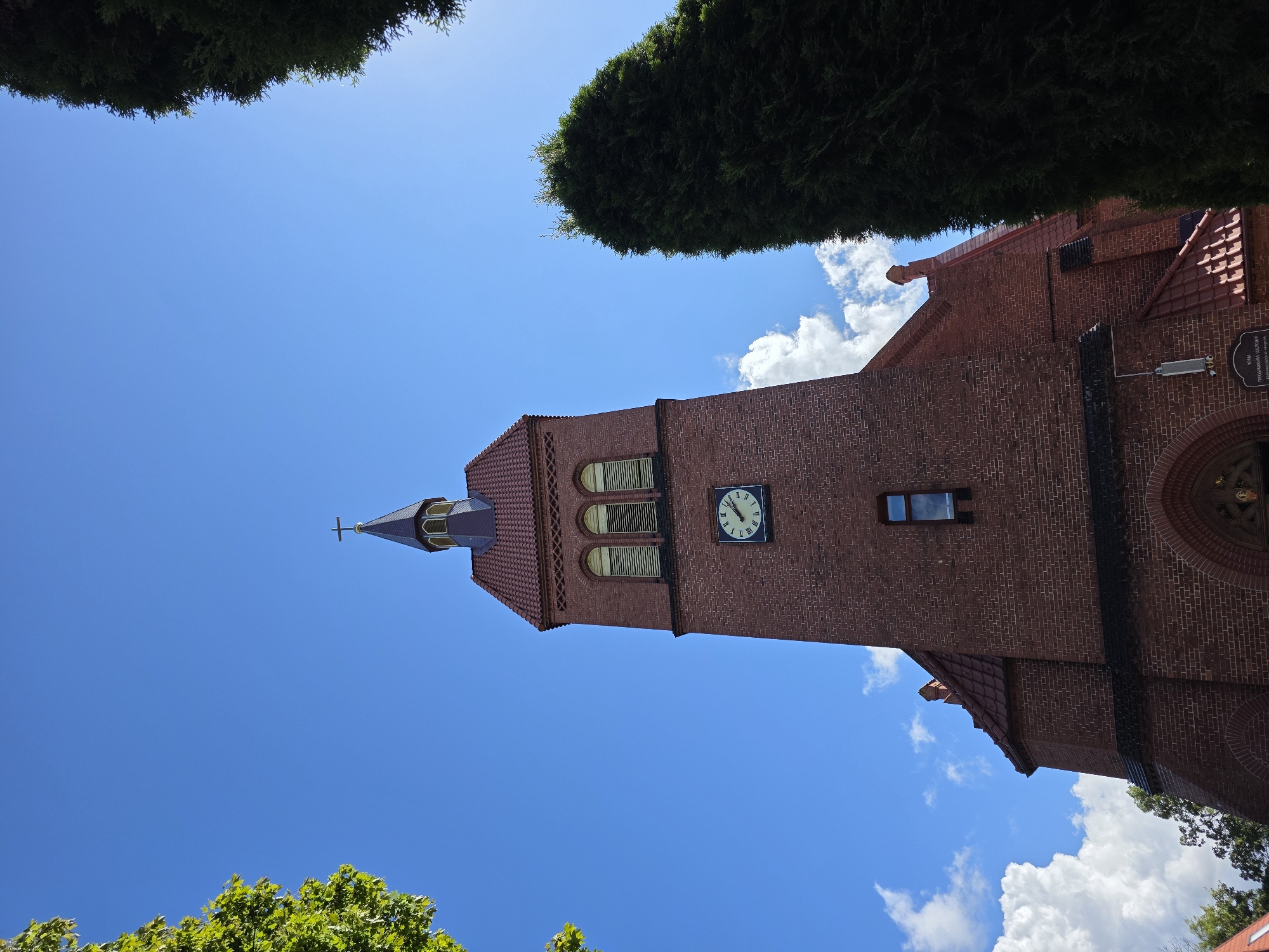 The image shows the exterior of a red brick church, likely located in Zelenogradsk, Russia, given the context. The church has a tall, prominent bell tower with a clock face visible. The clock has Roman numerals and the hands indicate approximately 10:10. Above the clock, arched windows are visible, and the tower is topped with a dark, pointed steeple surmounted by a cross. Red tile roofing is evident on the main building and the tower. The sky is a vibrant blue with scattered white clouds. Green foliage from trees frames the top and right sides of the image, partially obscuring the view of the church. A large, dark green evergreen tree stands to the right of the church tower. The bottom edge of the image shows the lower part of the church entrance archway and some text on a plaque. The scene is set during daylight, under a clear sky, suggesting pleasant weather. There are no people visible in the image.