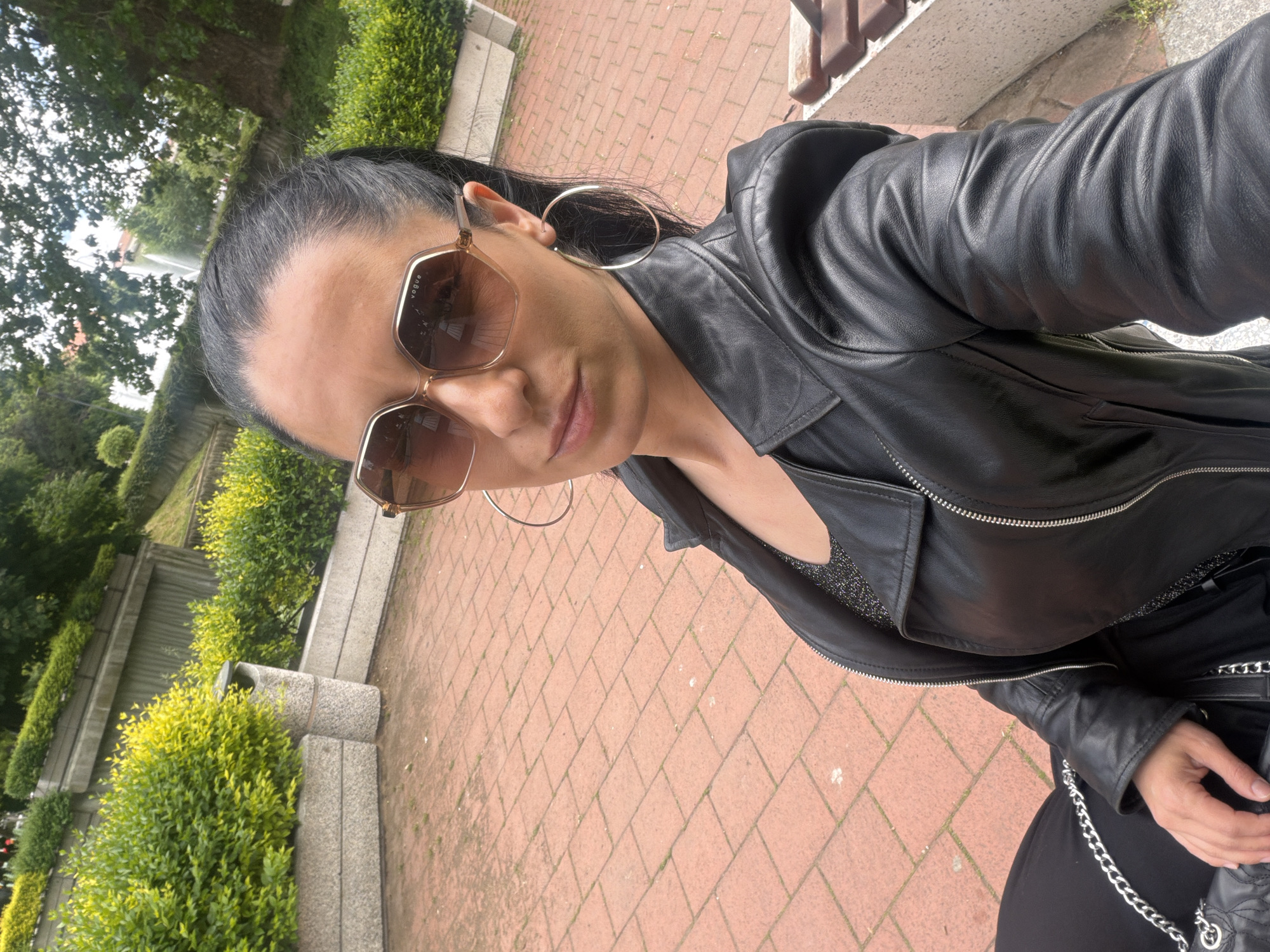 A woman is taking a selfie in Pleven, Bulgaria. She has dark hair pulled back into a ponytail, large hoop earrings, and sunglasses. She is wearing a black leather jacket over a dark top. The background shows a park with green bushes, trees, stone steps and a brick walkway. There is a fountain visible in the distance.