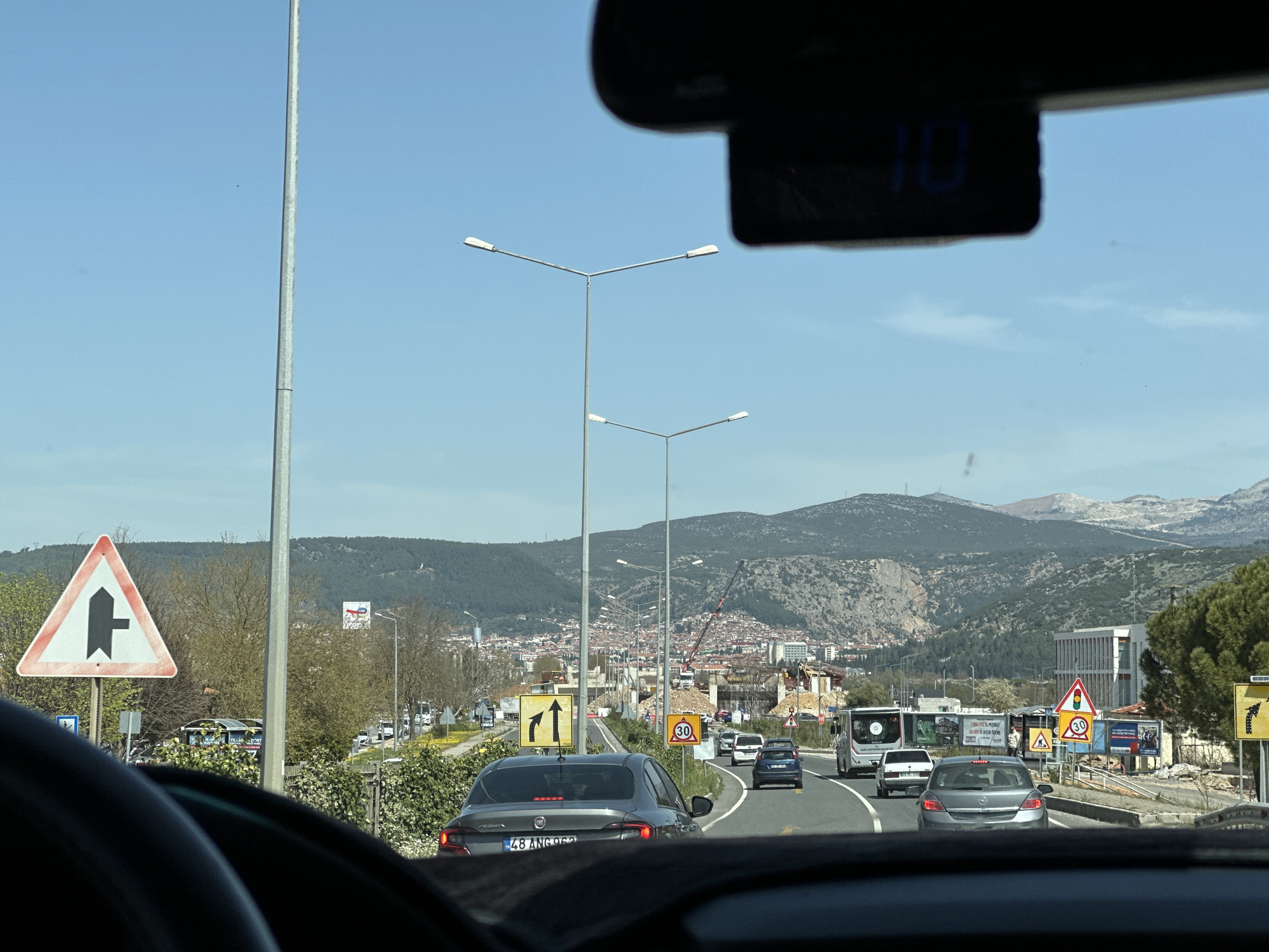 The image depicts a view from inside a car driving on a road in Menteşe, Turkey. The car is one of several in traffic on a multi-lane road. A gray sedan with license plate "48 ANG 962" is directly ahead. Road signs are visible on both sides of the road, including a triangular sign warning of a side road joining from the left and a sign indicating a speed limit of 30 km/h. A city with a mix of low-rise and taller buildings lies in the distance, backed by mountains. Overhead streetlights line the road. The sky is clear and blue. A small digital device is attached to the rearview mirror, displaying the number "10". A crane is working on construction of a tall building in the distance.