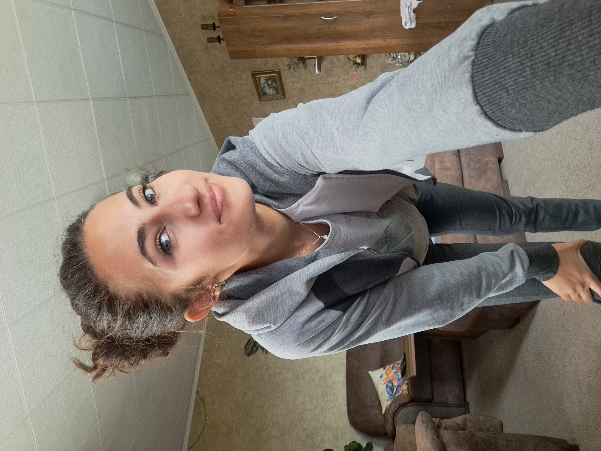 A young woman is taking a selfie indoors. She is wearing a grey hooded sweatshirt over a white shirt, and dark grey pants. Her hair is pulled back. In the background is a wall with square tiles, a wooden cabinet, a framed picture, a couch with a decorative pillow, and a plant. The location is Unknown, Unknown.
