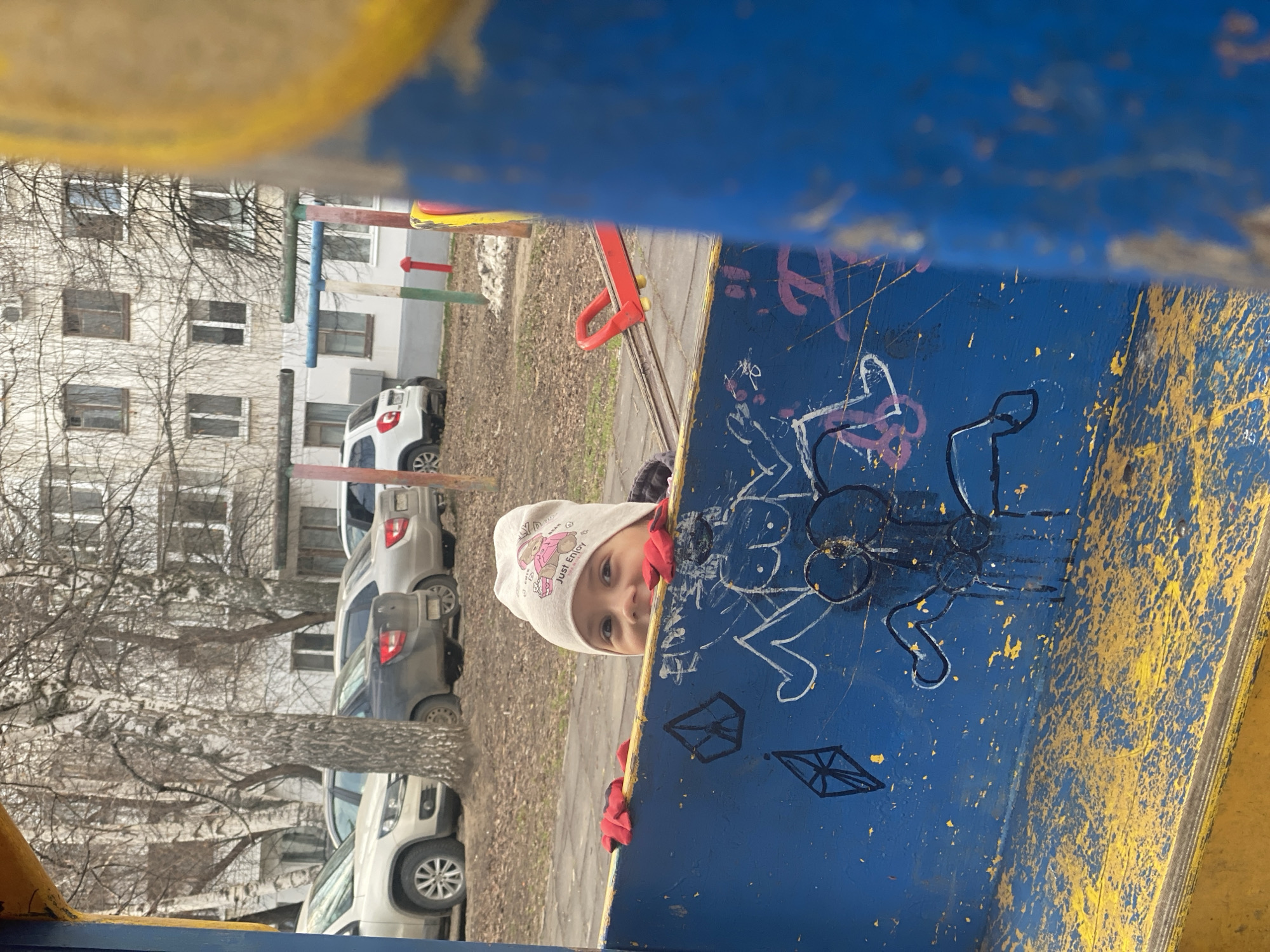 A young child peers out from behind a section of a blue-painted playground structure in Chaikovsky, Russia. The child, wearing a light-colored hat with a cartoon motif and red gloves, looks directly at the camera with one eye visible. The playground structure is marked with graffiti, including a depiction of a figure and other abstract designs in white and black. In the background, a row of parked cars is visible along with a multi-story building, devoid of foliage, indicating a colder season. The scene suggests an urban environment, likely a residential area.