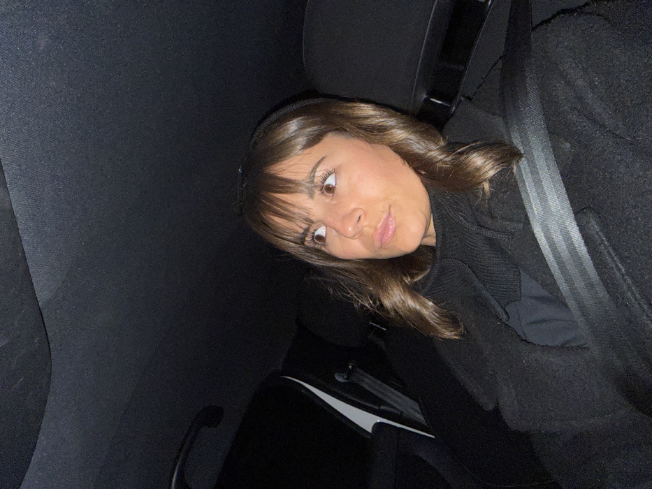 A young woman with brown hair and bangs glances at the camera, seated in a car. She wears a dark coat and a seatbelt is fastened across her chest. The car's interior is dark, with a black seat and ceiling. A white panel and a door handle are visible in the background. The image is angled, giving a tilted perspective.