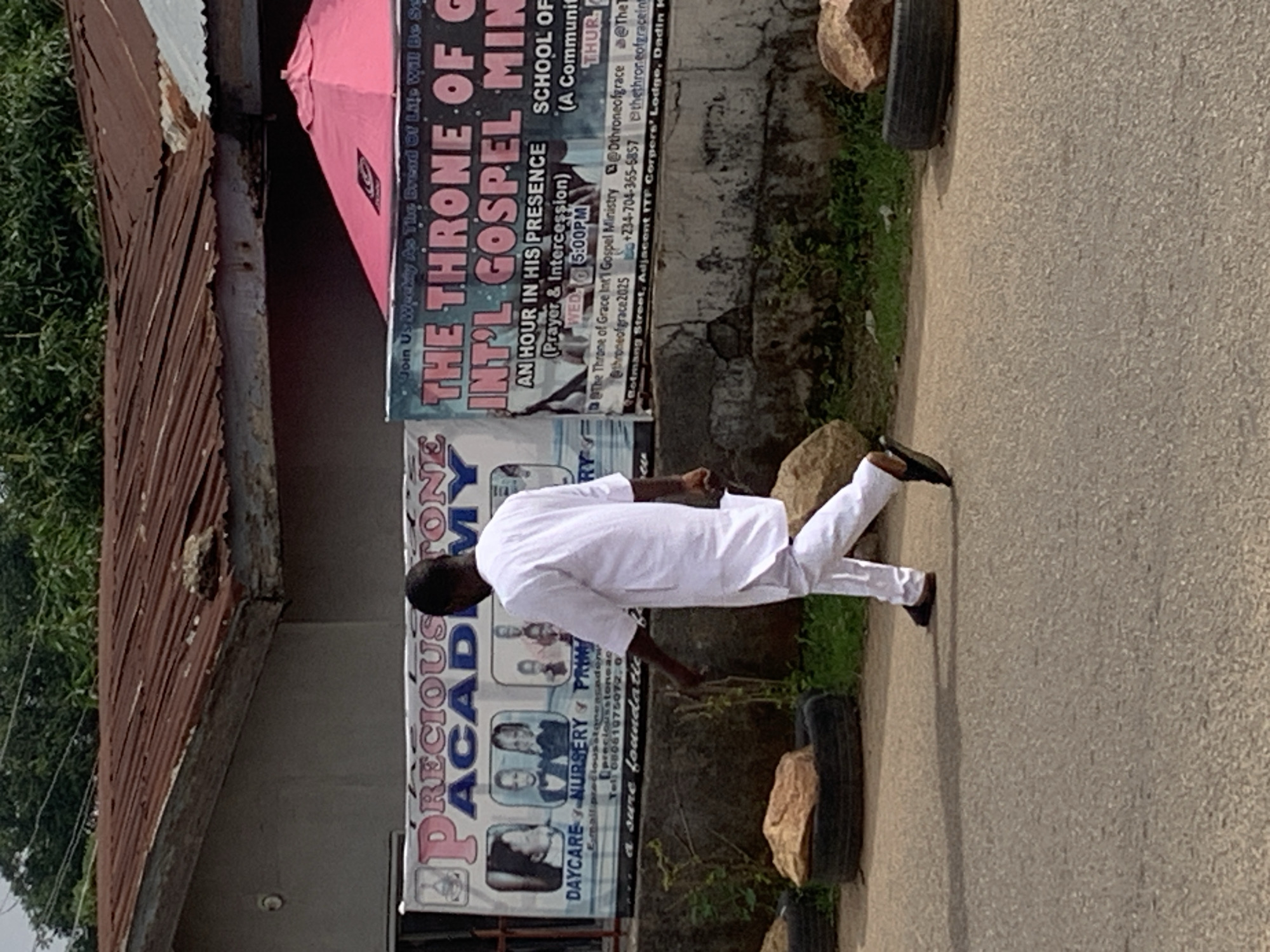 This outdoor image, taken in bright daylight and rotated 90 degrees clockwise from its original orientation, depicts a street scene in what appears to be Jos, Nigeria. The central figure is a person, likely a man, dressed in a traditional white tunic and trousers, walking along a paved path.
To the left of the path, a building with a rusty, corrugated iron roof is partially visible, alongside green foliage. Adjacent to the path, two large banners are prominently displayed against a wall. The banner closer to the viewer advertises "PRECIOUS ACADEMY" for "DAYCARE / NURSERY / PRIMARY" education, showing pictures of children and listing a contact number (08061075072). The second, larger banner promotes "THE THRONE OF GRACE INT'L GOSPEL MINISTRY," inviting people to "AN HOUR IN HIS PRESENCE (Prayer & Intercession)" on "WED. 5:00 PM," and mentioning a "SCHOOL OF" session on "THUR." This banner also provides social media handles (@Dthroneofgrace, @thethroneofgraceint, @throneofgrace2025) and a phone number (+234-704-365-6857), specifying the location as "Bolmang Street, Adjacent ITF Corpers' Lodge, Dadin K."
Behind these banners, a pink umbrella is partially visible. The area next to the path features patches of green grass, rocks, and dark-colored tires, possibly used as planters or barriers. The scene captures a mix of commercial and institutional elements in an urban or semi-urban setting.