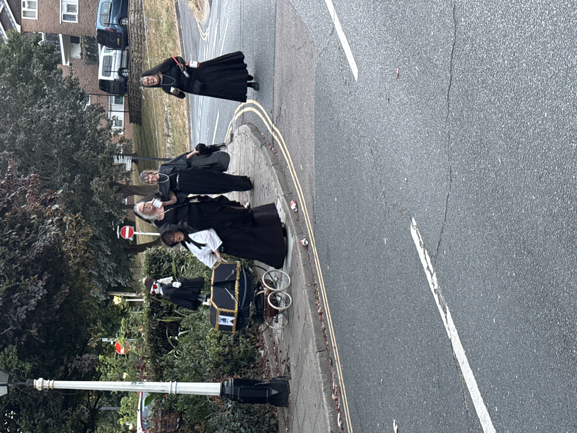 Four individuals, three women and one child dressed in historical Victorian-era clothing, are seen on a sidewalk next to a street. One woman is pushing a vintage-style baby carriage. The scene is set in St Leonards, United Kingdom. The background includes trees, houses, and vehicles. The day appears to be overcast.