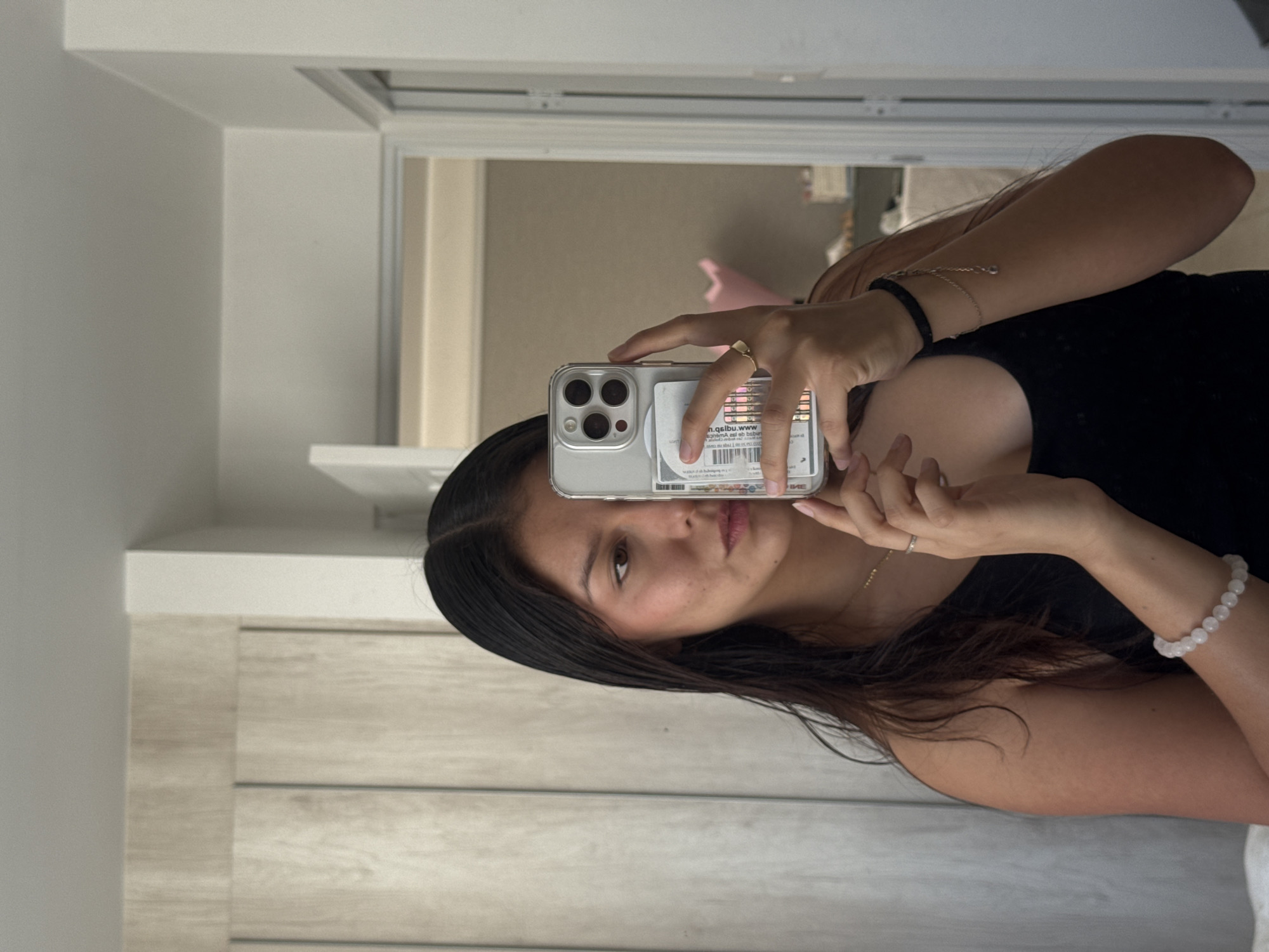 A young woman is captured in a mirror selfie, holding a silver-colored smartphone with a clear case. Her face is partially visible behind the phone, with a neutral expression as she looks towards the camera. She has long, dark hair that falls over her shoulders and wears a dark top.
Her hands are prominent, holding the phone. On her right hand, a simple gold-colored ring is visible on her ring finger. Her left wrist is adorned with a black band and a thin, silver-toned chain bracelet. On her right wrist, she wears a light-colored beaded bracelet, possibly made of rose quartz.
The background of the mirror reflection shows an indoor setting with light-colored walls and what appears to be light wooden paneling or furniture on the lower half. In the upper part of the image, a doorway or window frame is visible. There's a small, white card or sticker inside the phone's clear case, displaying faint, mirrored, and largely illegible text, including what appears to be "IME" and a fragment like "n.qspu.ww." The overall lighting suggests an indoor environment.