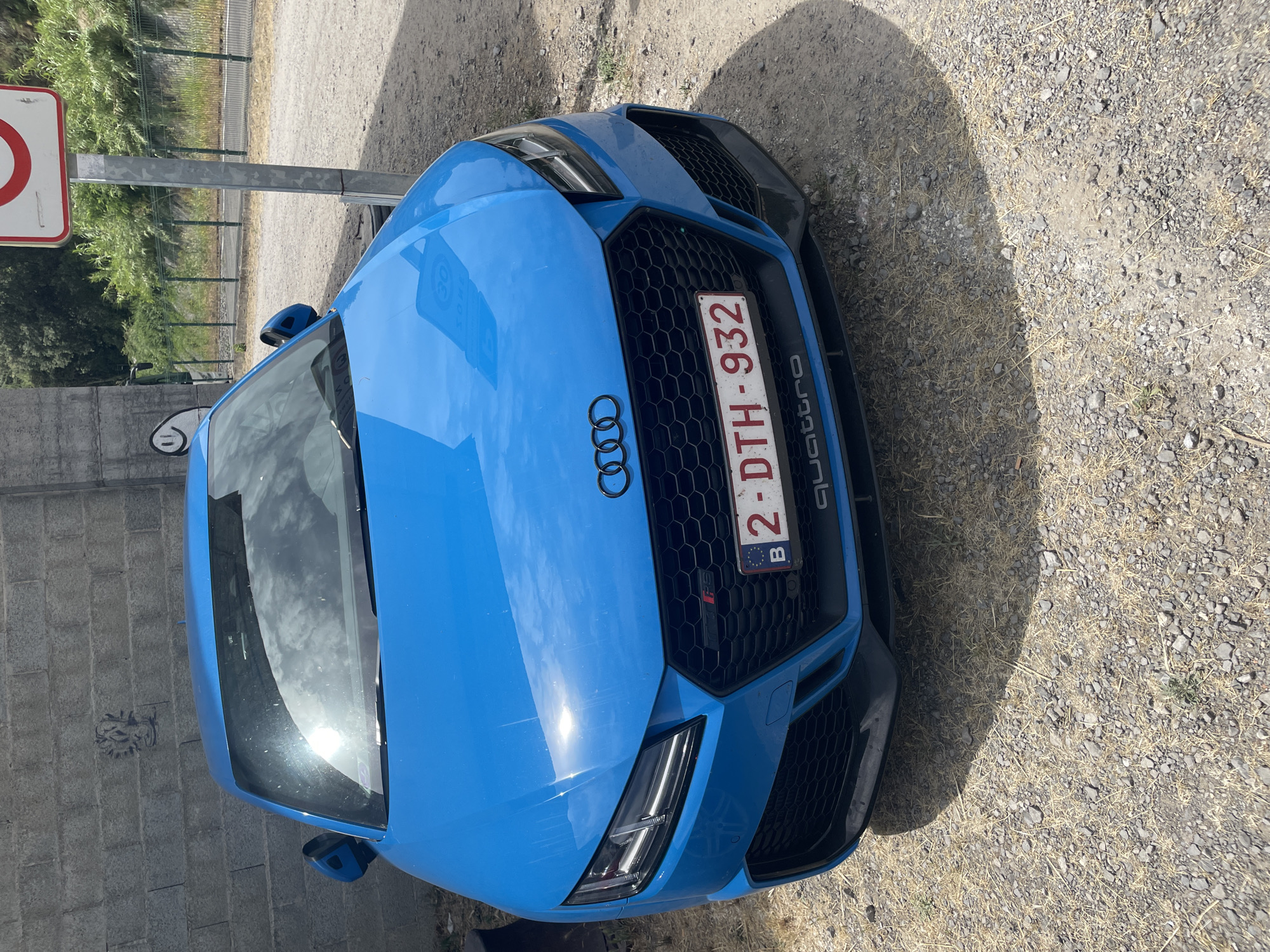 A blue Audi TT RS is parked on a gravel surface next to a grey brick wall. The car has a black honeycomb grille with the Audi rings logo in black above it. A Belgian license plate, "B 2-DTH-932", is mounted on the front of the car. The word "quattro" is printed in silver below the license plate. The car's headlights are on. In the background, there is a metal pole with a circular sign on top, and green foliage beyond a fence. The sun is shining, casting a shadow from the car onto the ground. There is graffiti on the brick wall.