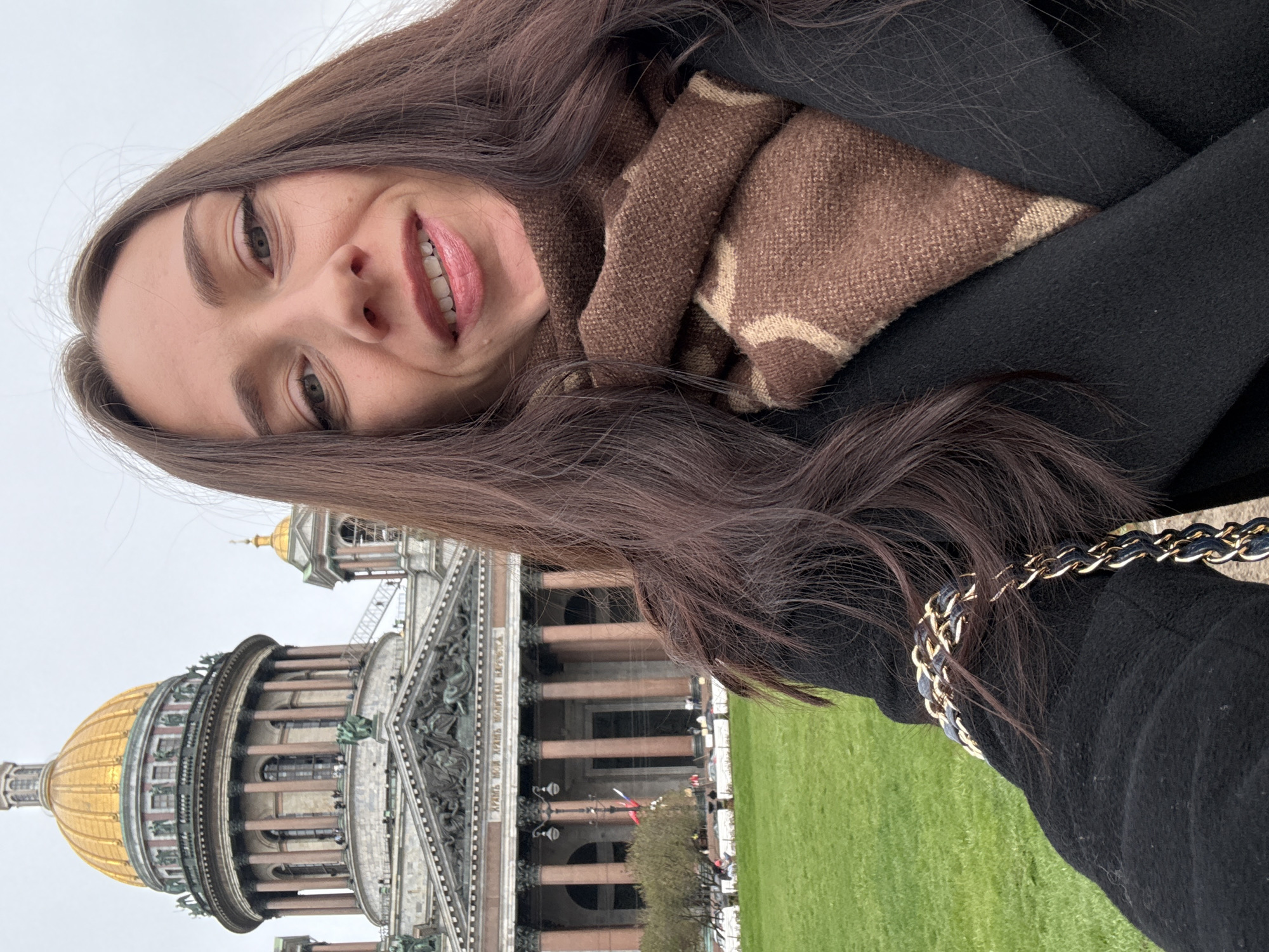A selfie of a woman with long brown hair, wearing a black coat and brown patterned scarf, with the Saint Isaac's Cathedral in Saint Petersburg, Russia, as a background. The woman is smiling and looking at the camera. A gold-colored dome of the cathedral is partially visible. There is a lawn to the side of the cathedral. The picture is taken at a tilted angle. A chain accessory is also visible.