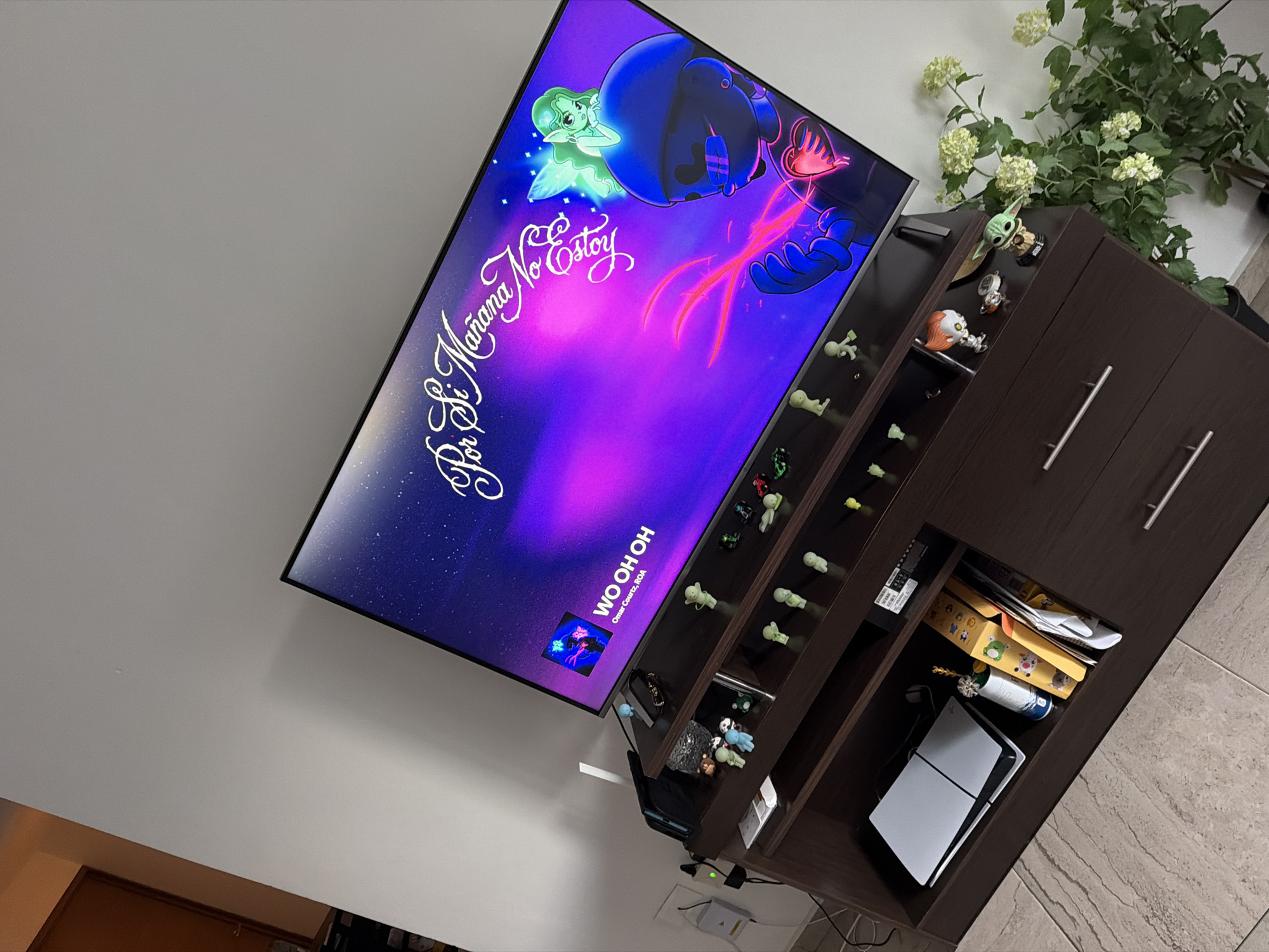 An indoor scene in San Andrés Cholula, Mexico, showcases a wall-mounted flat-screen television displaying vibrant digital artwork. The TV screen features a purple and blue gradient background with animated characters: a glowing green fairy-like figure on the left and a larger, dark blue entity with red glowing accents on the right. Visible text on the screen reads "Por Si Mañana No Estoy" in a cursive white font, with "WO OH OH" and "Omar Coutz, ROA" below it, indicating a music video or album art.
To the right of the television stands a dark brown entertainment unit or cabinet with multiple open shelves. The top of the cabinet holds a green plant with clusters of light-colored, small flowers. The shelves are adorned with numerous small collectible figurines, including a Baby Yoda (Grogu) Funko Pop, a Pennywise (IT) Funko Pop, an astronaut figure, and several light green and blue glow-in-the-dark-style figures. The bottom-right section of the cabinet houses a white PlayStation 5 console, a decorative foil-wrapped bottle, and a stack of papers and a yellow box. A remote control is also visible on one of the shelves. The wall behind the setup is plain light gray or white, and the floor is covered in light-colored tiles. The scene suggests a personalized entertainment area, likely a living room, captured from a slightly elevated, tilted perspective.