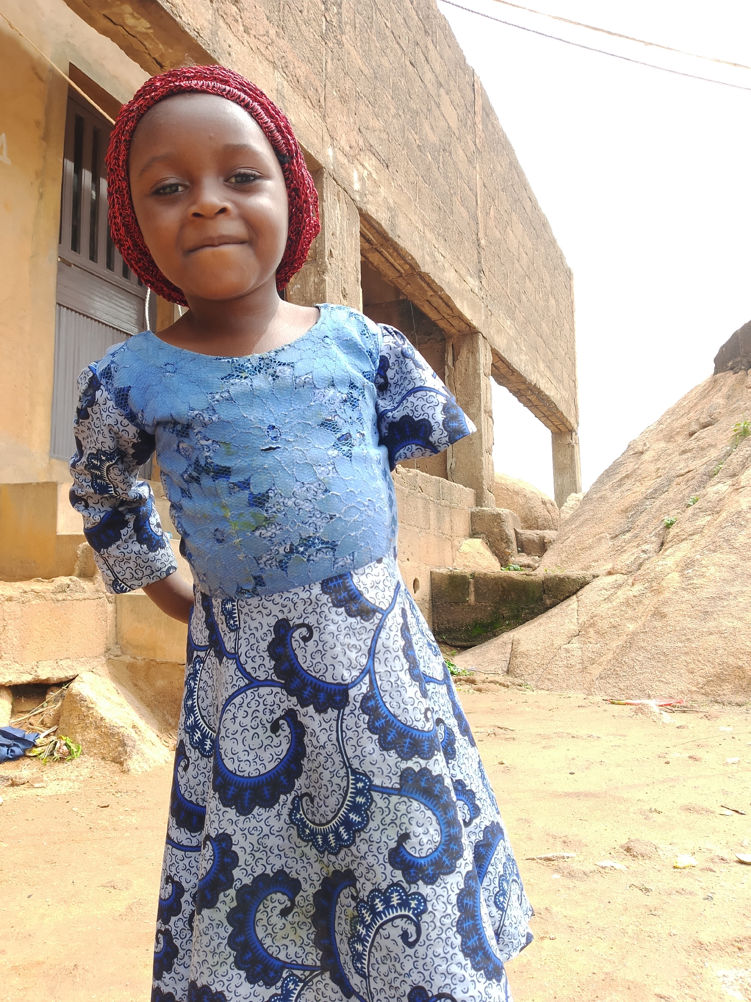 A young Black girl stands outside in front of a building. She is wearing a blue and white patterned dress with a red head covering. She has her hands behind her back. The building appears to be made of concrete blocks and brick, and there is a large rock formation behind it. The ground is covered in dirt and sand. The sky is bright and overcast.