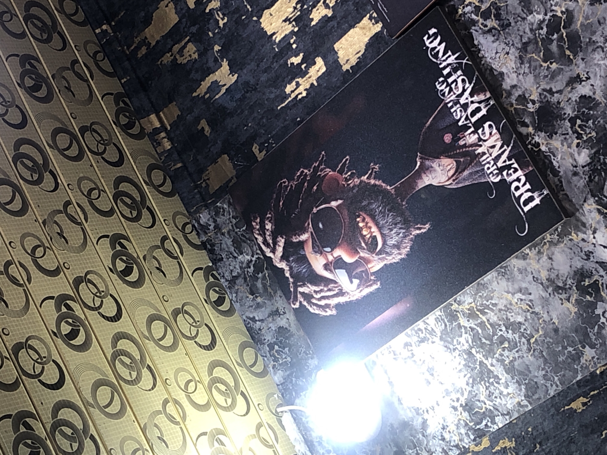 The image shows a close-up, slightly angled view of two rectangular prints laid on a dark, textured surface with gold accents. The print in the foreground features a cartoonish character with dreadlocks, sunglasses, and a prominent smile with visible teeth. The character is wearing a dark garment with what appears to be tattoos on its neck. Below the character, white text reads "GRILL FLASHING DREAMS DASHING". A bright, blown-out light source is visible on the left side of the image, casting a strong glare. The background consists of a ceiling with a repeating pattern of concentric circles in black and gold. The overall impression is a display of artwork in an indoor setting.