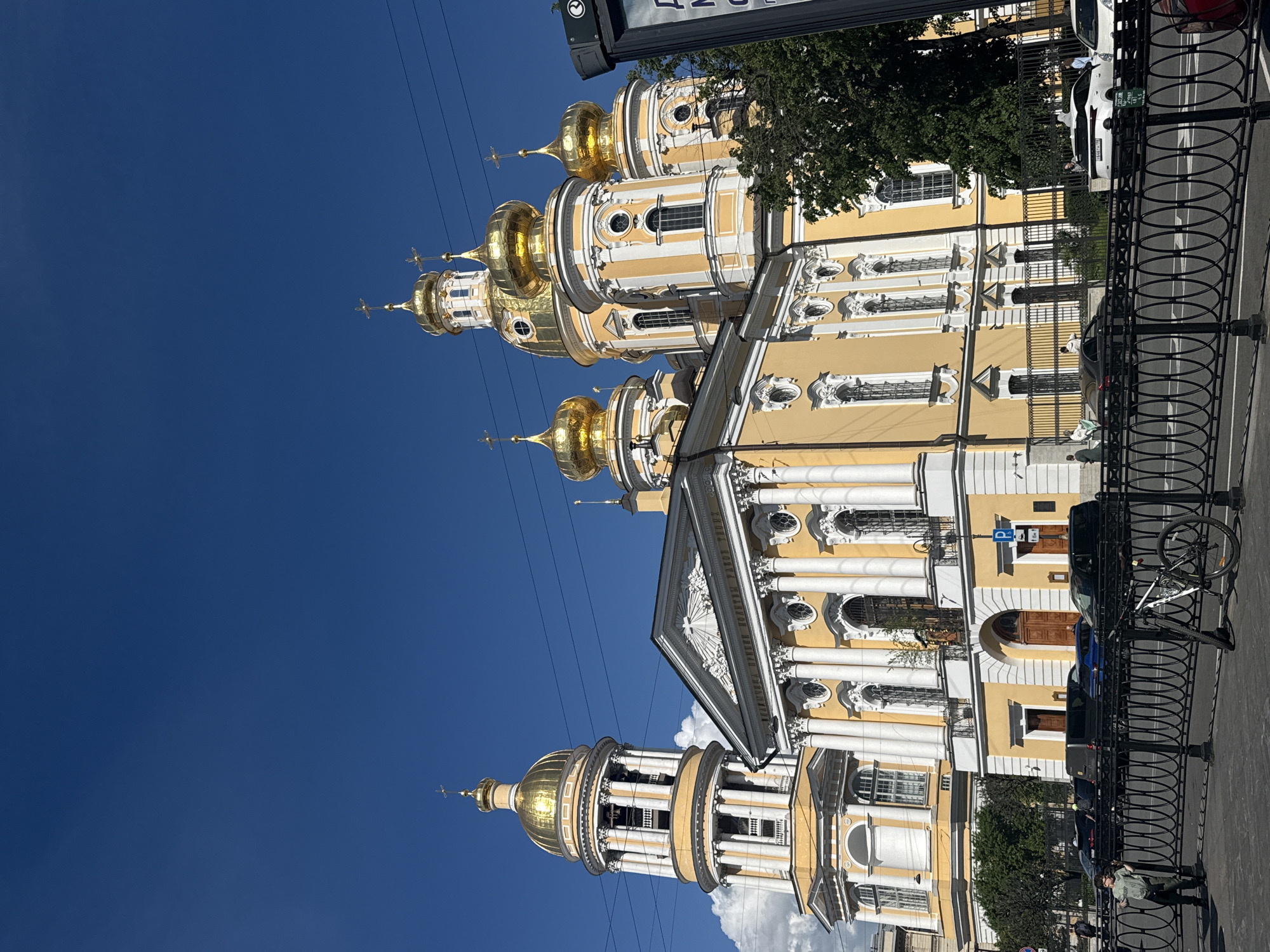 This image captures the exterior of the magnificent St. Nicholas Naval Cathedral (Nikolsky Cathedral) in Saint Petersburg, Russia, under a clear, deep blue sky. The grand cathedral features a vibrant yellow facade adorned with intricate white classical details, including columns, decorative window frames, and cornices. Multiple golden onion-shaped domes, each crowned with an Orthodox cross, reflect the bright sunlight. Inscriptions in Cyrillic script are visible on the pediment of one section of the building.
The scene depicts a bright, sunny day with a few scattered white clouds. In the foreground, a black metal fence with ornate circular patterns separates the cathedral's grounds from a city street. Overhead power lines traverse the upper portion of the sky.
Several cars are parked along the street, including a white car, a dark vehicle, and a blue car. A bicycle leans against the fence. A pedestrian is visible walking on the sidewalk near the fence. Green trees provide natural elements to the urban setting behind the fence. A small, partially obscured sign with white text, possibly including Russian words like "УЛ" (Street), is also visible in the upper right corner. The photograph conveys a vibrant urban environment showcasing a significant architectural landmark.
