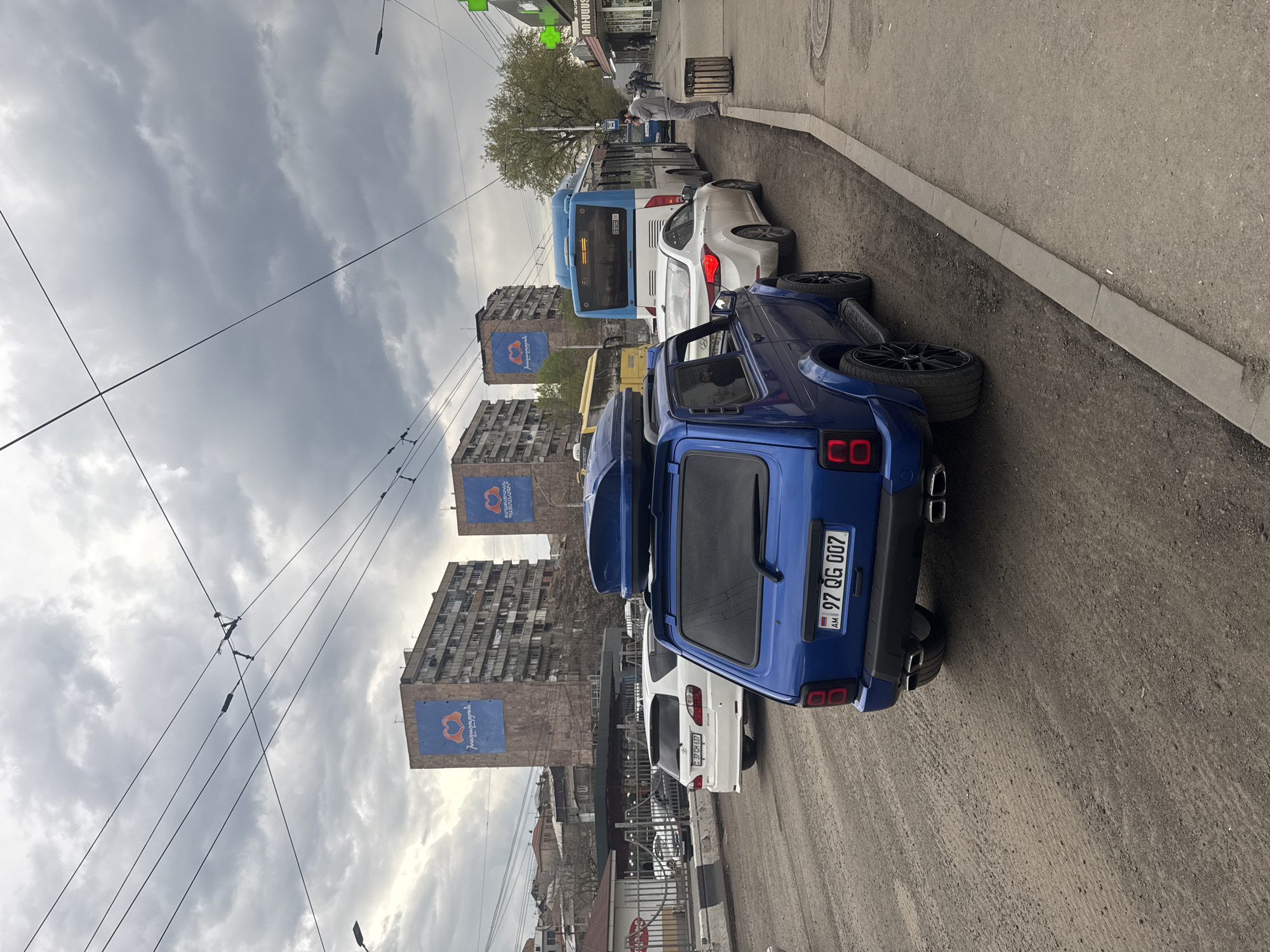 The image depicts a street scene in Yerevan, Armenia, characterized by a row of parked vehicles along the curb. A blue custom SUV with license plate "97 QG 007 AM" occupies the foreground, directly behind a white sedan. Further down the street, a blue and white bus is visible. Buildings with large blue banners featuring a heart-shaped design are prominently displayed in the background. Overhead power lines stretch across the cloudy sky. A person in a gray suit is standing on the sidewalk at the corner of the image with his back facing towards the camera.