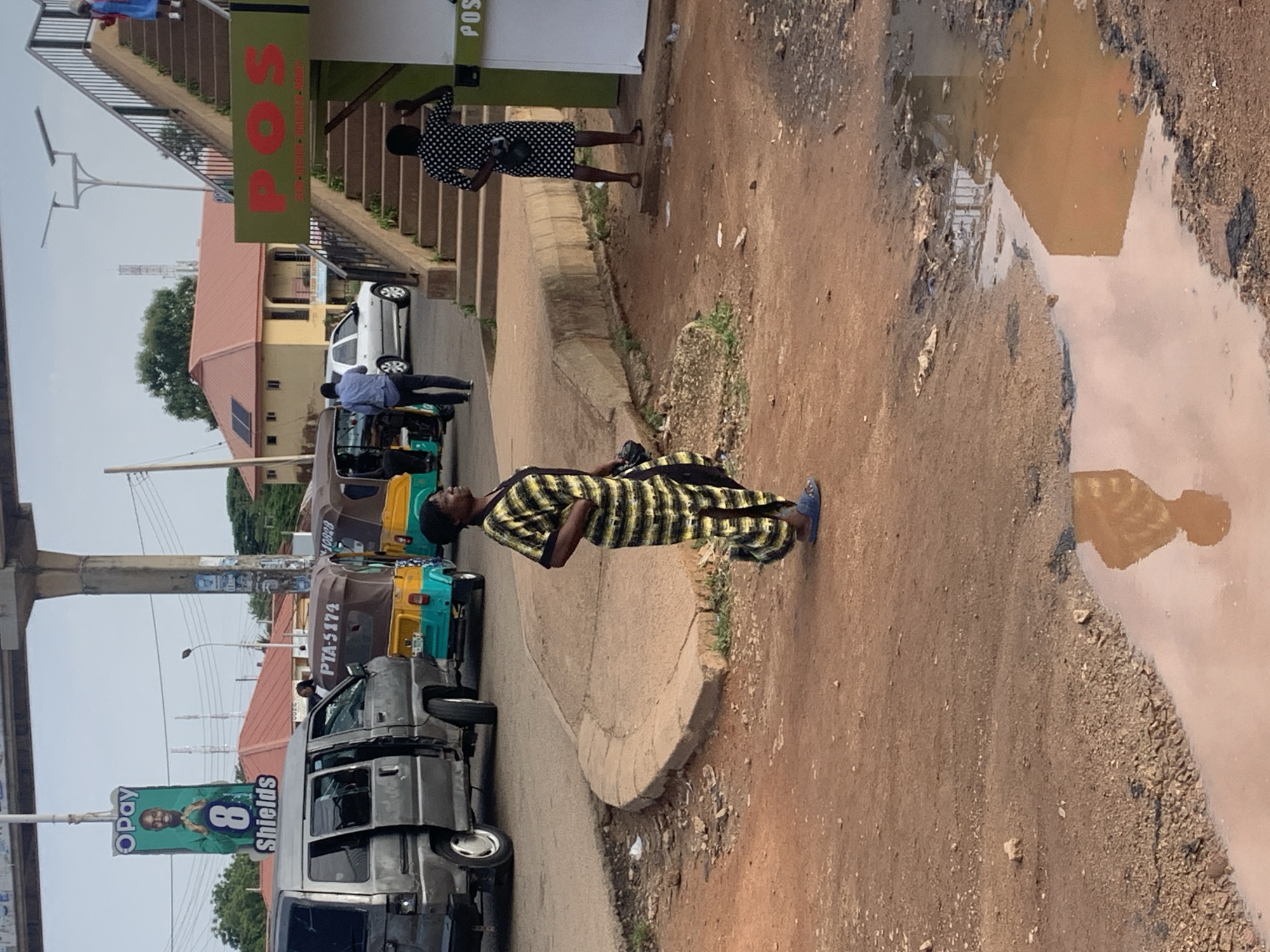The image depicts a street scene in Gura Top, Nigeria, with a focus on pedestrian activity and vehicles. In the foreground, a woman in a yellow and black striped dress stands on a dirt path, looking to her right. Adjacent to her, a large puddle reflects the surrounding environment. Behind her, several vehicles are present, including a gray van and three colorful auto-rickshaws. A pedestrian bridge with stairs is visible in the background, with two individuals ascending it. A sign for "POS" is prominently displayed near the top of the stairs. An OPay advertisement featuring a man is mounted on a pole. The overall environment appears to be a bustling urban area with a mix of infrastructure and daily life.
