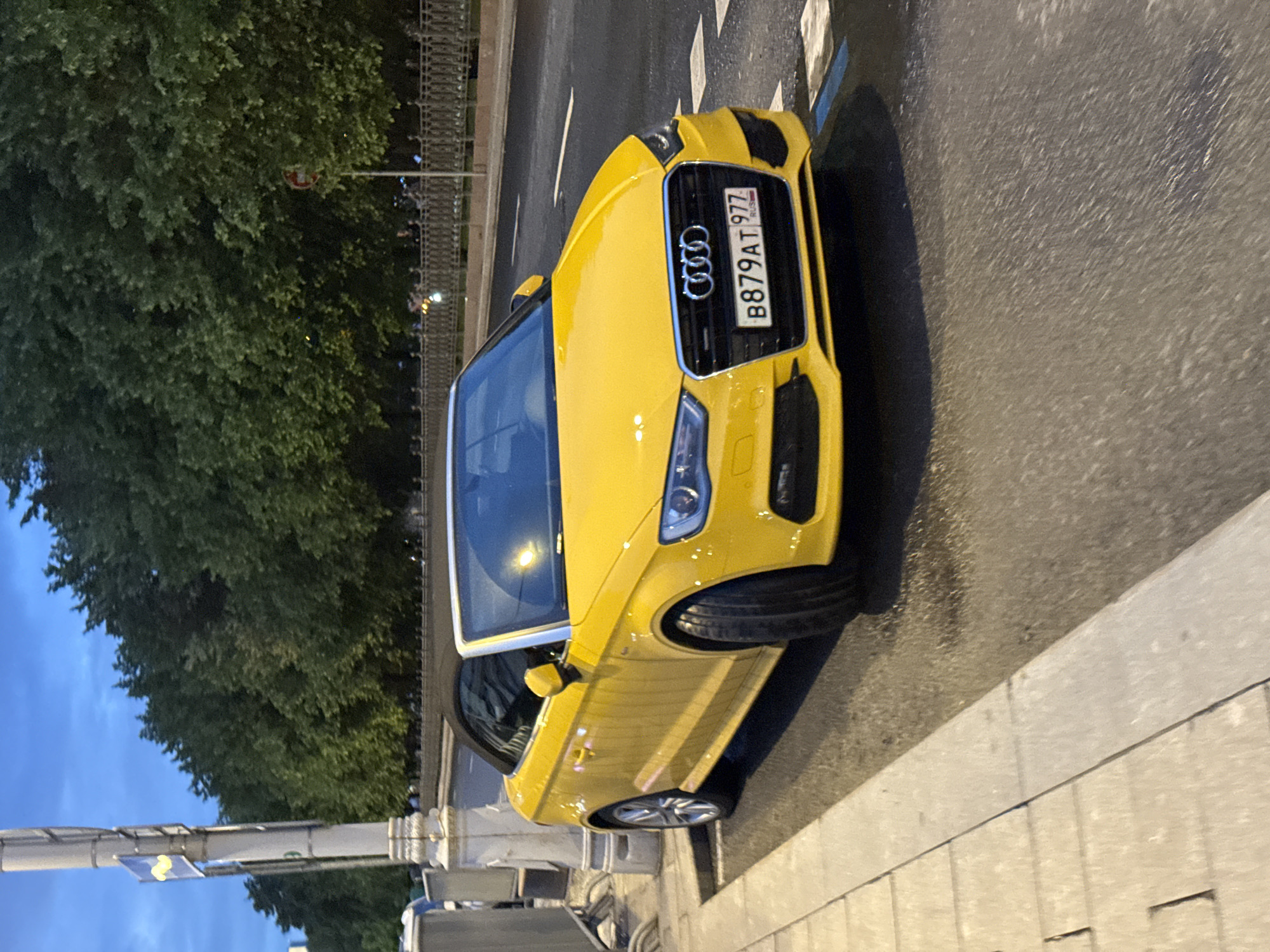 A yellow Audi A3 Cabriolet, bearing Russian license plate B879AT 777, is parked on a city street in Moscow, Russia. The car is facing the camera. A tree line is visible to the left, and a street light is also visible. The surface of the street appears wet.