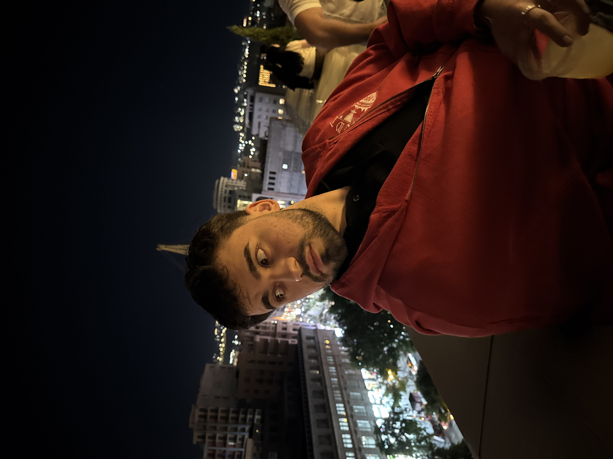 A tilted, medium shot depicts a man with dark hair and a beard, wearing a partially unzipped red hoodie over a black shirt, looking at the camera. He appears to be outdoors at night, with city buildings and lights visible in the background. Another person, wearing a white shirt, is partially visible above and to the right of the man. The man holds a translucent plastic cup. The image appears to have been taken in Yerevan, Armenia, based on the architectural style of the buildings and the general urban environment. A construction crane is slightly visible on the left side of the frame.