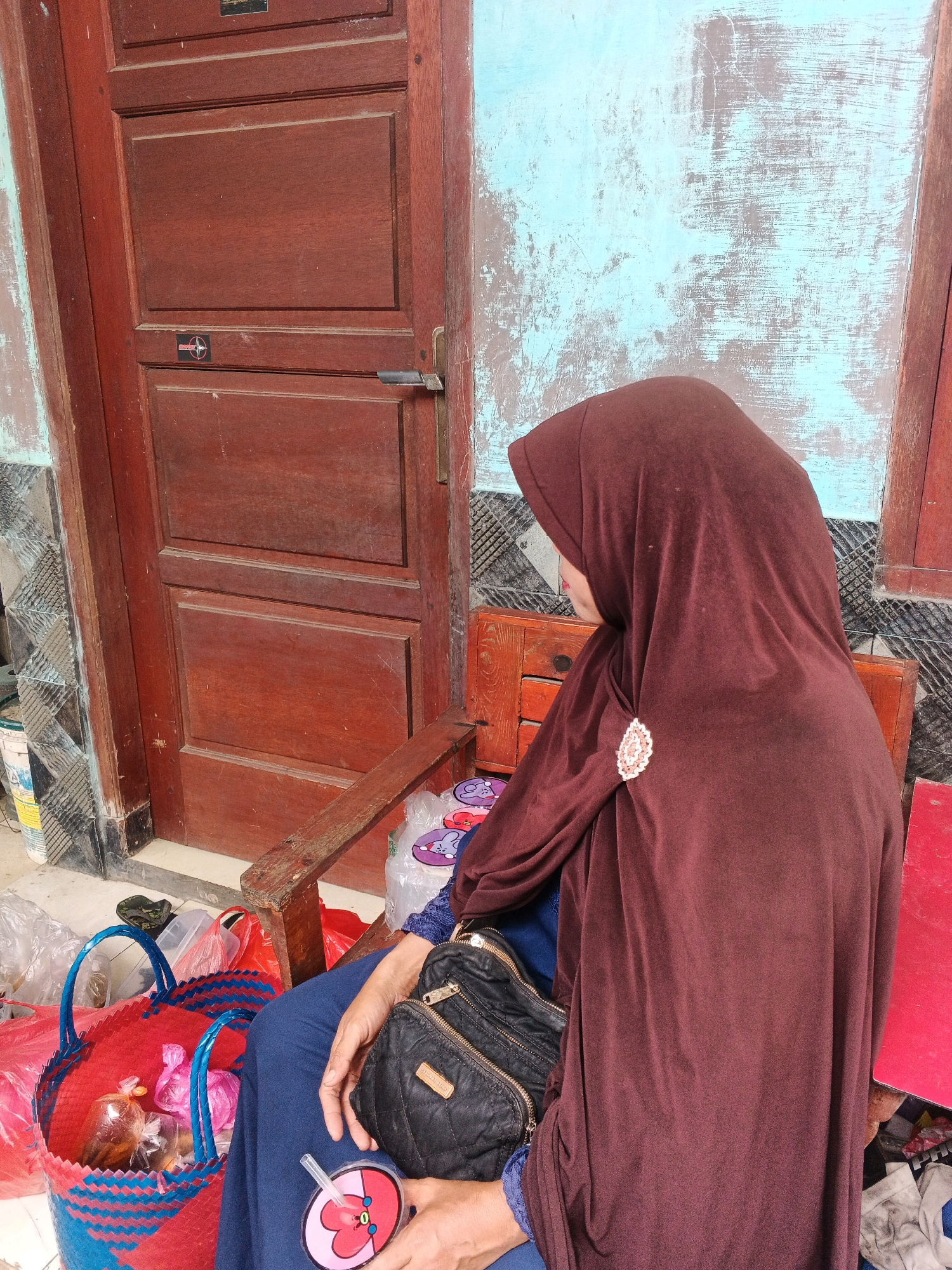 A woman wearing a brown hijab and blue clothing sits on a wooden chair outside. She is holding a black purse and a circular drink with a straw. In front of her, a woven red and blue basket contains several plastic-wrapped items. Behind her is a wooden door with multiple panels and a metallic handle. The wall to the right of the door is painted a distressed light blue. The ground is paved with concrete and there are other bags and objects around. The image appears to be taken in the city of Kadu, Indonesia, suggesting an outdoor setting in front of a dwelling.