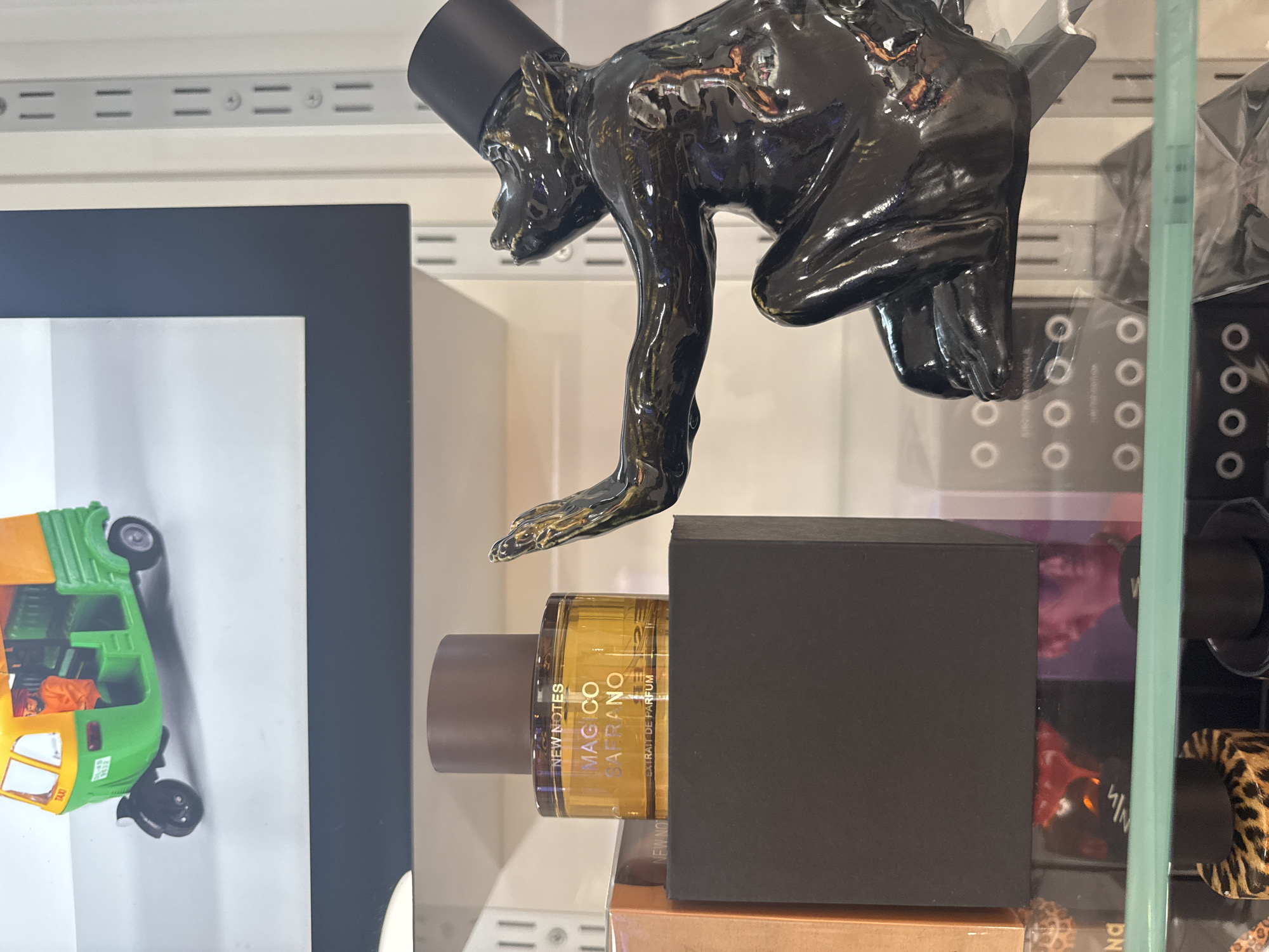 The image depicts a shelf display in a retail setting, likely a perfume or gift shop. In the foreground, a glass bottle of "MAGICO SAFRANO" extrait de parfum by NEW NOTES is prominently displayed on a black box. To the right, a glossy black statue of a monkey wearing a top hat is positioned, its arm extended as if reaching for something. In the background, a miniature yellow and green auto-rickshaw is visible on an upper shelf. The shelves are made of metal and white material, with a light blue shelf edge in the foreground. The overall lighting suggests an indoor environment, possibly a store with artificial lighting. The location is identified as Germasogeia Municipality, Cyprus, indicating a commercial establishment within that area. There are no people visible in the image.