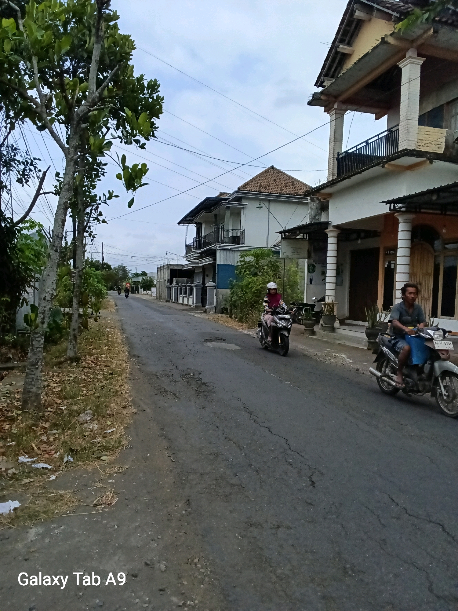 The image depicts a street scene in Kelutan, Indonesia, under an overcast sky. A paved road with visible cracks runs through the middle of the frame, flanked by vegetation and buildings. On the right side of the road, a two-story house with a covered porch and balconies stands out, featuring white columns and a light yellow facade. Further down the street, another building with a tiled roof and white walls is visible, with a blue and grey sectional building to its right.
Two individuals are on motorcycles, moving along the road. The person in the foreground, on the right, is an adult male with short dark hair and a casual grey shirt, riding a grey motorcycle with a blue bag strapped to the front. The second motorcyclist, further up the road on the left, is an adult female wearing a white helmet and a patterned headscarf with a pinkish hue, riding a dark-colored motorcycle. Trees with sparse green foliage line the left side of the road, with some fallen leaves scattered on the verge. The overall impression is of a quiet, residential street in a tropical or subtropical climate. The text "Galaxy Tab A9" is watermarked in the lower left corner of the image.