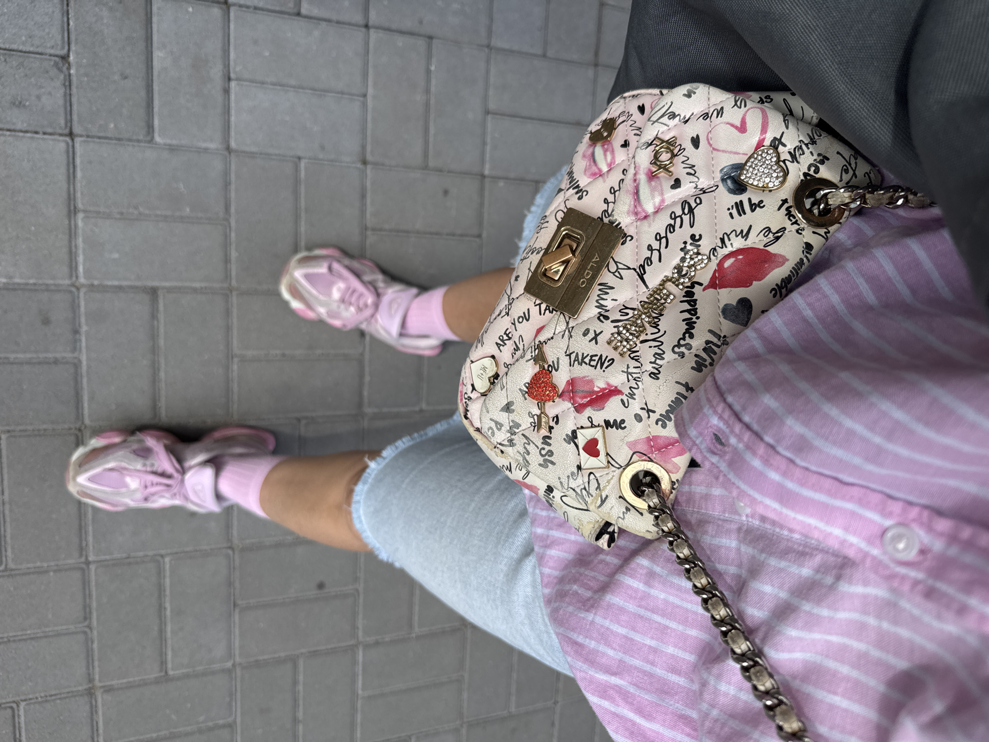 An overhead, ground-level shot captures a person's legs, adorned in pink socks and chunky pink sneakers, standing on a grey brick pathway. The individual is wearing light blue denim shorts and a pink and white striped button-down shirt. A small, quilted crossbody bag with a white background and a pink graphic print, featuring hearts, lips, and cursive text, rests on the person's lap, secured by a silver chain strap. The bag has a gold clasp and is embellished with various charms, including a red heart with an arrow, a small envelope with a red heart, and sparkling heart-shaped studs. The scene is static and depicts a casual outdoor setting, possibly a street or park. The specific location of Vsevolozhsk, Russia, cannot be determined from the visual information provided.