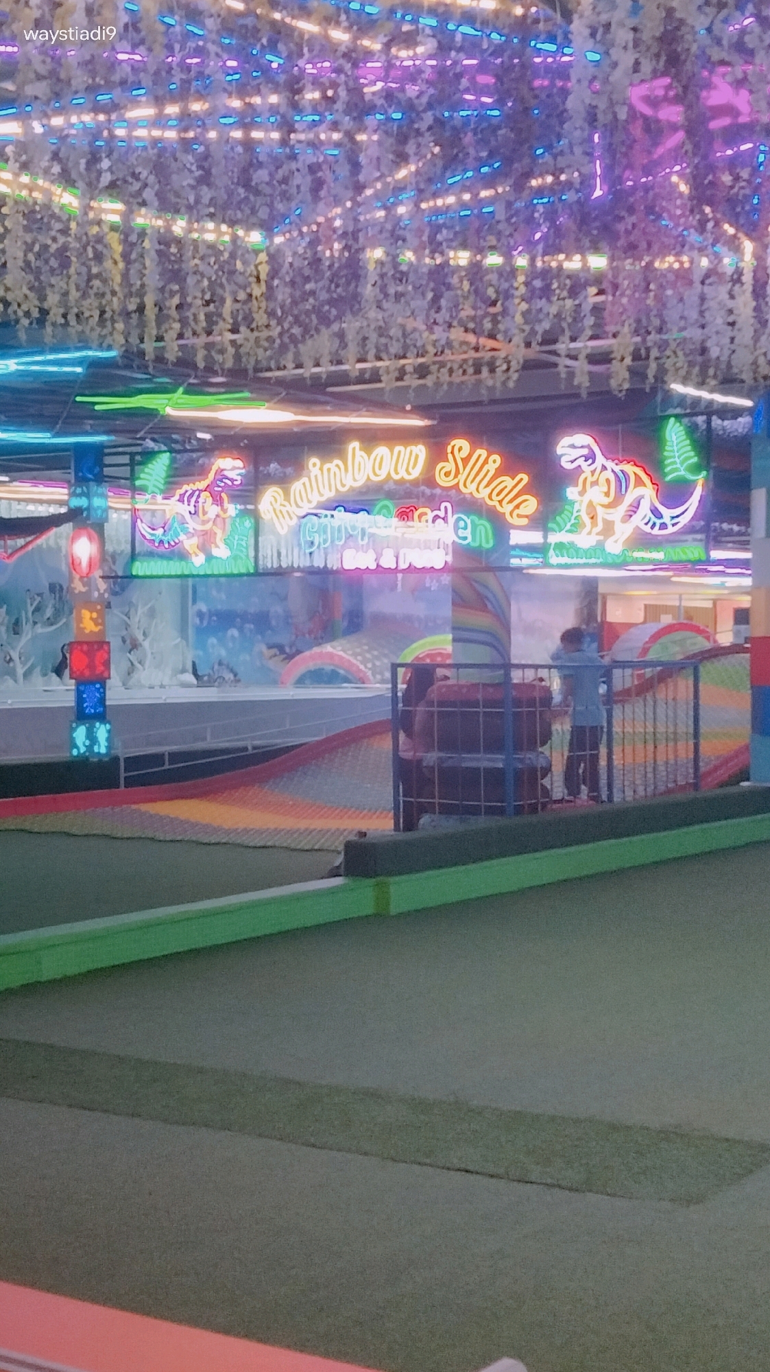 The image shows an indoor playground with neon lights and decorative greenery hanging from the ceiling. In the center, a sign reads "Rainbow Slide City Garden" in bright neon lettering, with neon dinosaur figures on either side. The playground features a colorful slide, a padded play area, and a net enclosure. A young person in a blue shirt and dark pants is visible near the net enclosure. The foreground consists of a green, carpeted area, possibly part of a mini-golf course. The overall impression is of a vibrant and playful indoor entertainment space. Based on the watermark "waystiadi9" and the common use of such neon-lit playgrounds in popular tourist areas, this location is likely in Bandung City, Indonesia, a city known for its numerous family-friendly attractions.