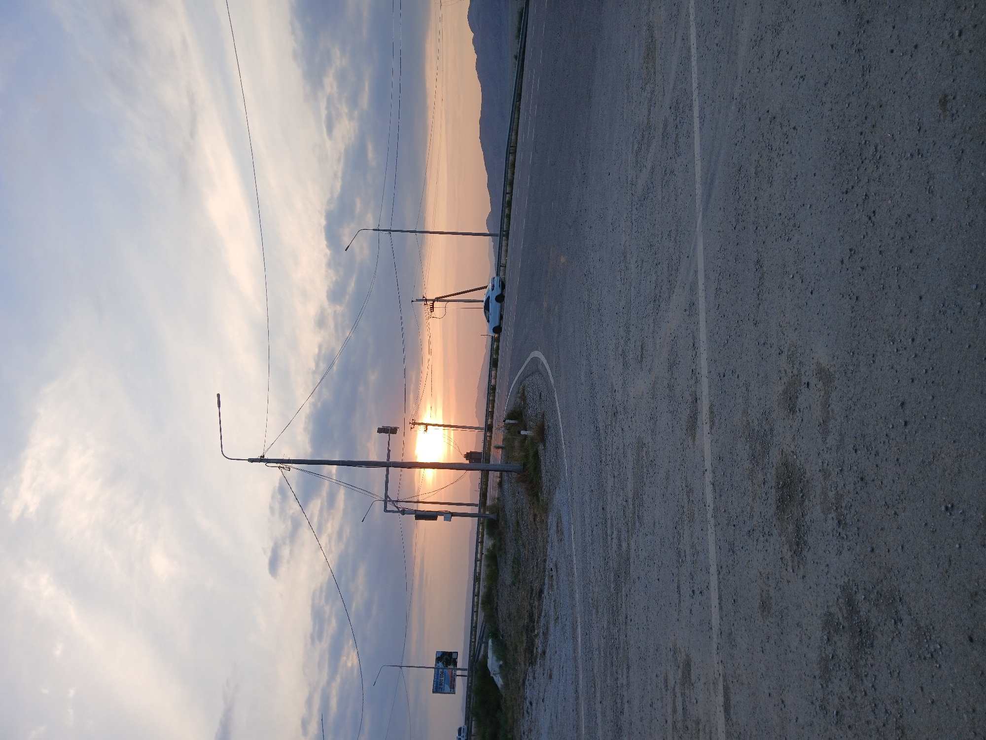 The image depicts a roadside scene at sunset in E'lmelikol, Russia. The sun is low in the sky, casting a warm, orange glow through the clouds and over a distant mountain range. The sky is filled with a mix of soft, wispy clouds and darker, more ominous ones.
The foreground is dominated by a paved road with a white lane marking. To the left of the road, there are several utility poles with wires stretching across the frame. A white car is parked on the side of the road near one of the poles. In the distance, more utility poles are visible, as well as some vegetation and what appears to be a billboard.
The overall mood of the image is serene and peaceful, with the natural beauty of the sunset contrasting with the man-made elements of the road and utility infrastructure. The time of day is clearly indicated by the sunset lighting. There is no visible text that can be clearly read.