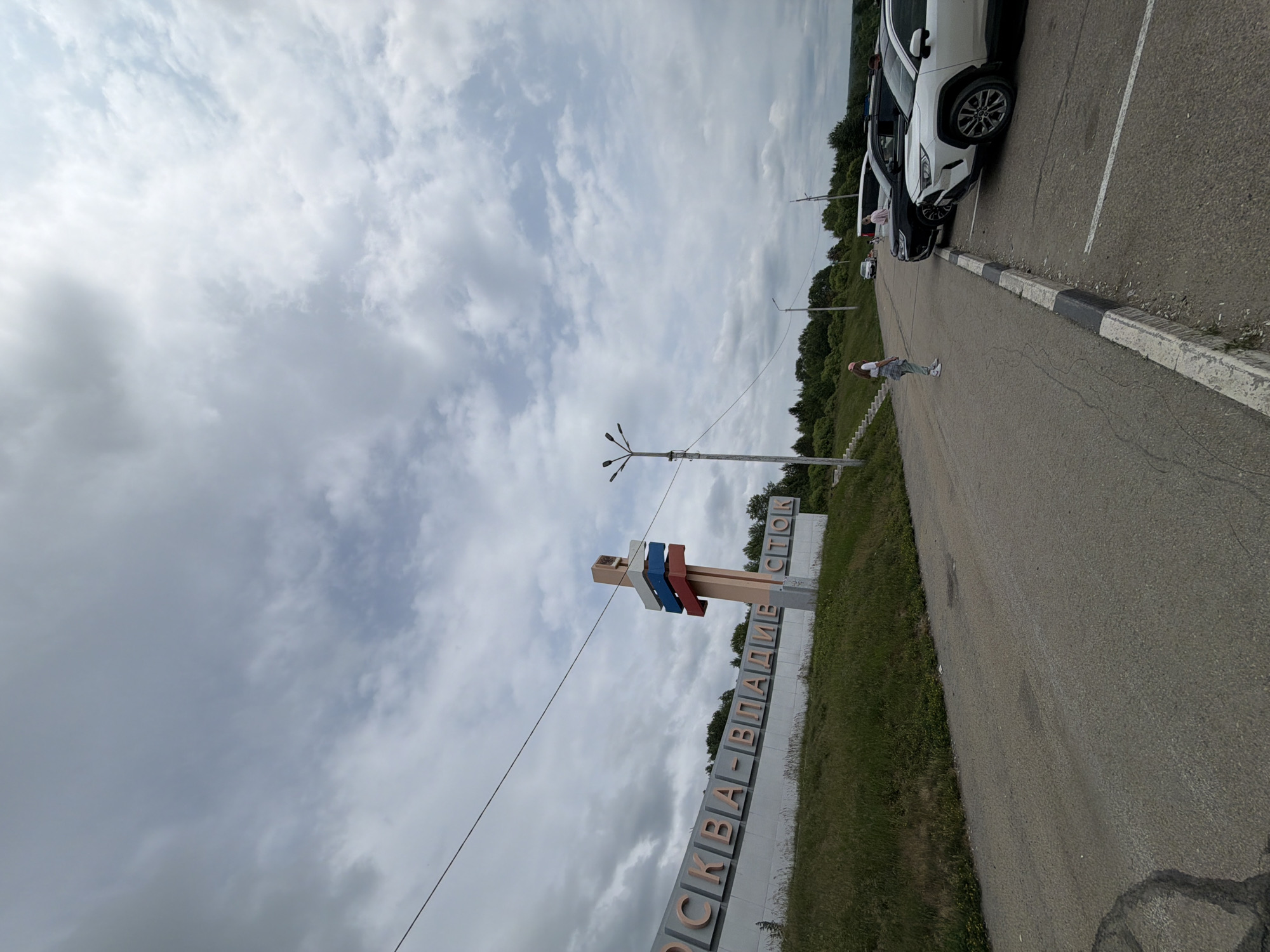 The image depicts a street scene in Nikolaevka, Russia, under a cloudy sky. A large, elevated sign with Cyrillic lettering reads "МОСКВА-ВЛАДИВОСТОК," indicating the route connecting Moscow and Vladivostok. A tall pole with three horizontal colored panels, resembling the Russian flag, stands next to the sign. Several cars are parked on the right side of the road, including a white SUV in the foreground. A young woman in a white shirt and light-colored pants stands on the side of the road, facing away from the camera. Another person is visible near the parked cars. The road surface appears to be asphalt, and a grassy embankment with trees lines the left side of the road, leading up to the sign. Streetlights are visible in the background.