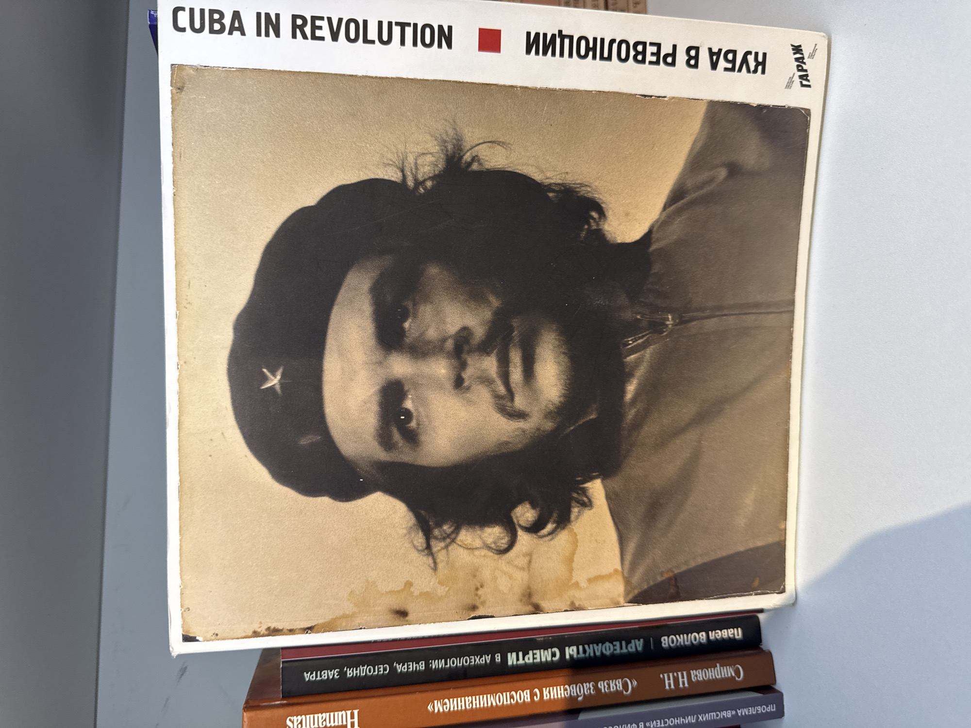The image shows a book titled "CUBA IN REVOLUTION" with the same title in Russian, "КУБА В РЕВОЛЮЦИИ," and a photograph of Che Guevara on the cover. The book sits on top of a stack of three other books, whose titles are partially visible. From top to bottom, they read: "Павел ВОЛКОВ | АРТЕФАКТЫ СМЕРТИ В АРХЕОЛОГИИ: ВЧЕРА, СЕГОДНЯ, ЗАВТРА", "Смирнова Н.Н. «Связь забвения с воспоминанием»", and "ПРОБЛЕМА «ВЫСШИХ ЛИЧНОСТЕЙ» В ФИЛО," with the word "Humanitas" also visible on the third book. The location in the city of Moscow, Russia is not explicitly stated or identifiable in this image.