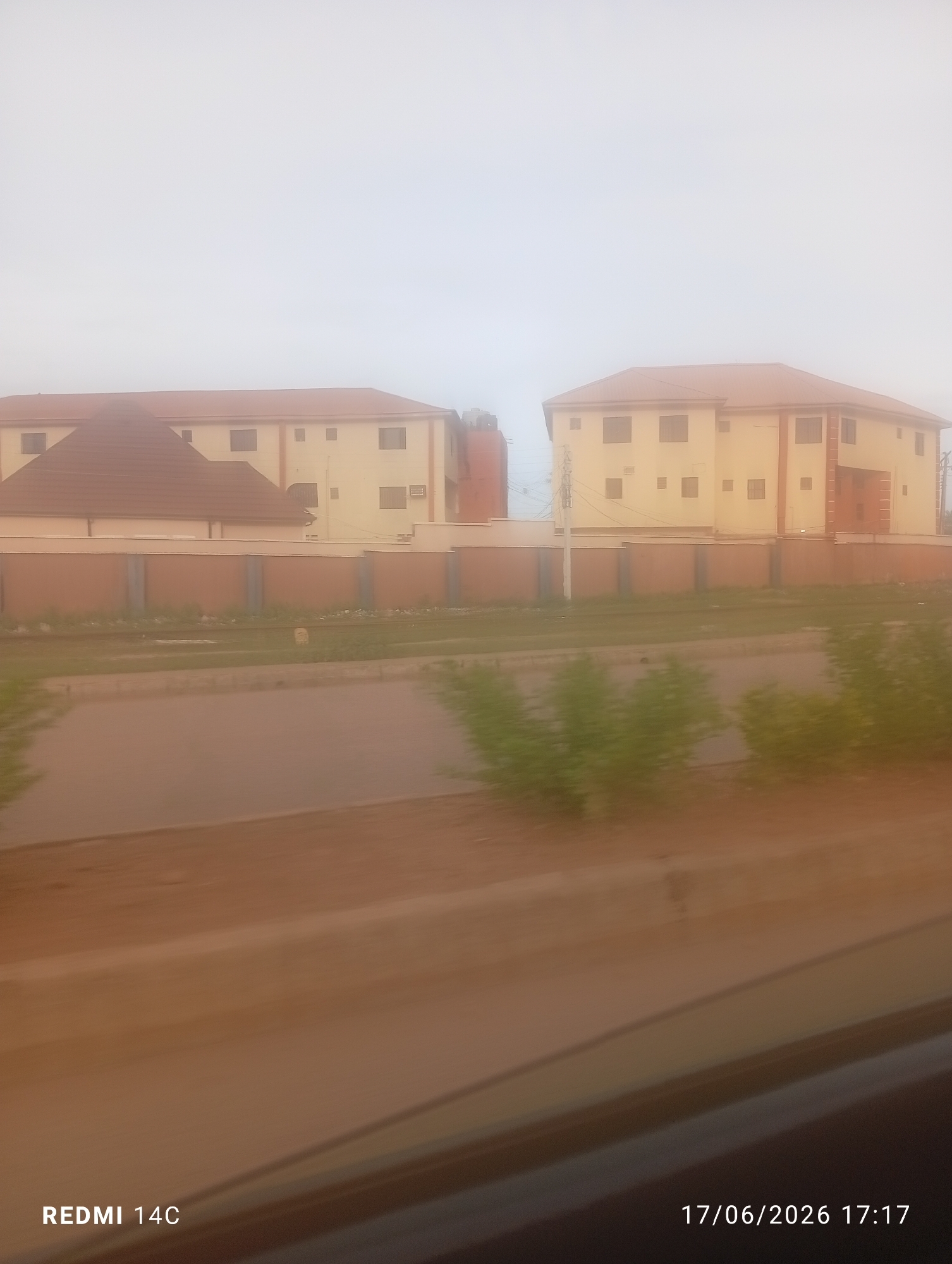 This is a photo of two buildings with reddish-brown roofs and cream-colored walls, fenced by a brown wall. The buildings have multiple windows and appear to be a hotel or apartment complex. In the foreground, there is a dirt road and some greenery. The sky is overcast, suggesting a cloudy day. The image seems to have been taken from a moving vehicle, as indicated by the blur in the foreground. The text "REDMI 14C" and "17/06/2026 17:17" are visible at the bottom left and right corners, respectively, indicating the phone model used and the date and time the photo was taken. The location is identified as Bukuru, Nigeria. No people are visible in the image.