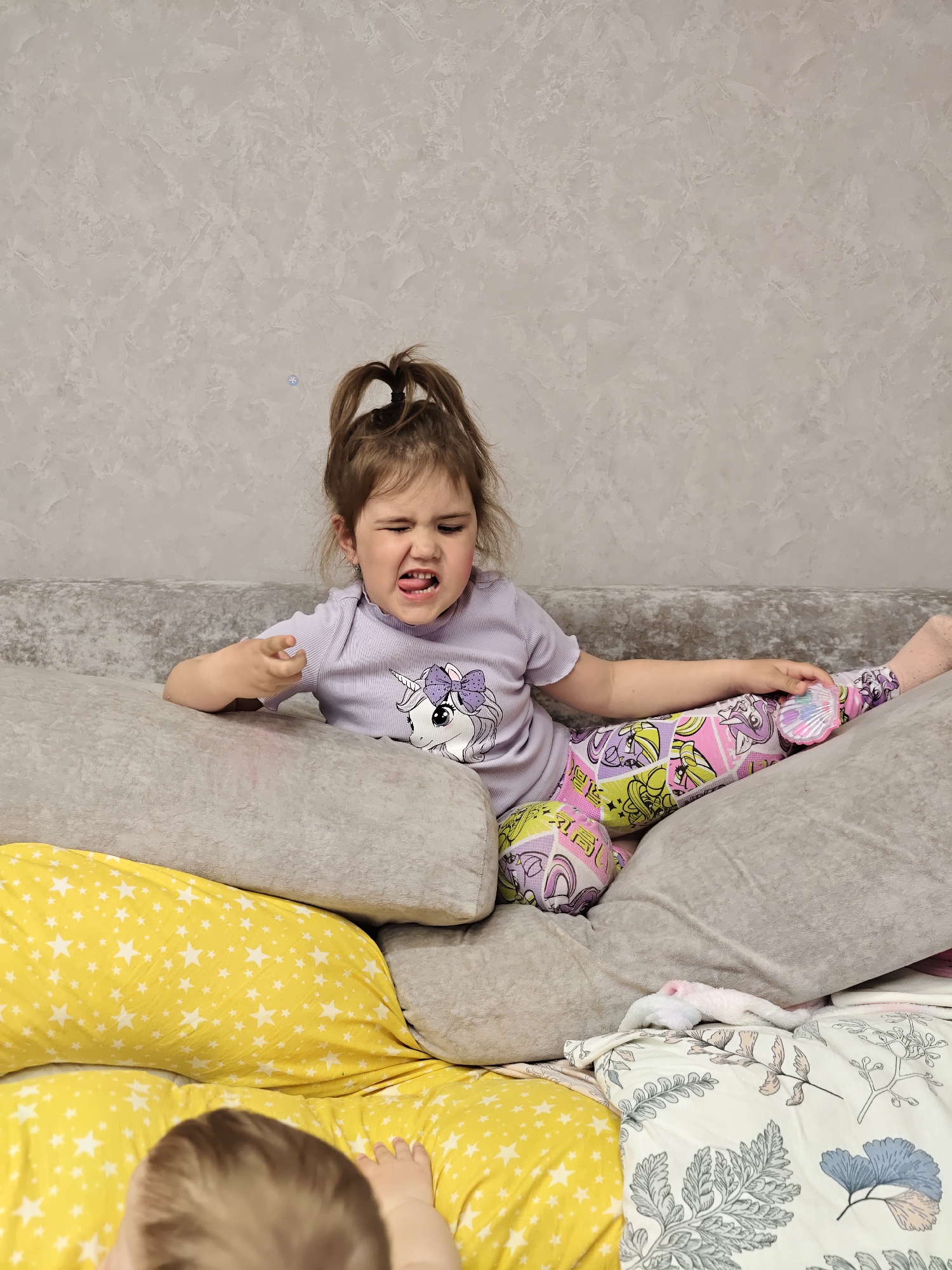 A young girl with brown hair pulled up in a ponytail sits on a pile of pillows and blankets. She wears a light purple shirt with a unicorn design and colorful patterned pants. She is sticking her tongue out and making a face. The pillows are gray and yellow with white stars. A baby's head and hand are visible in the lower left corner. The background is a textured gray wall. The location is Chaikovsky, Russia.