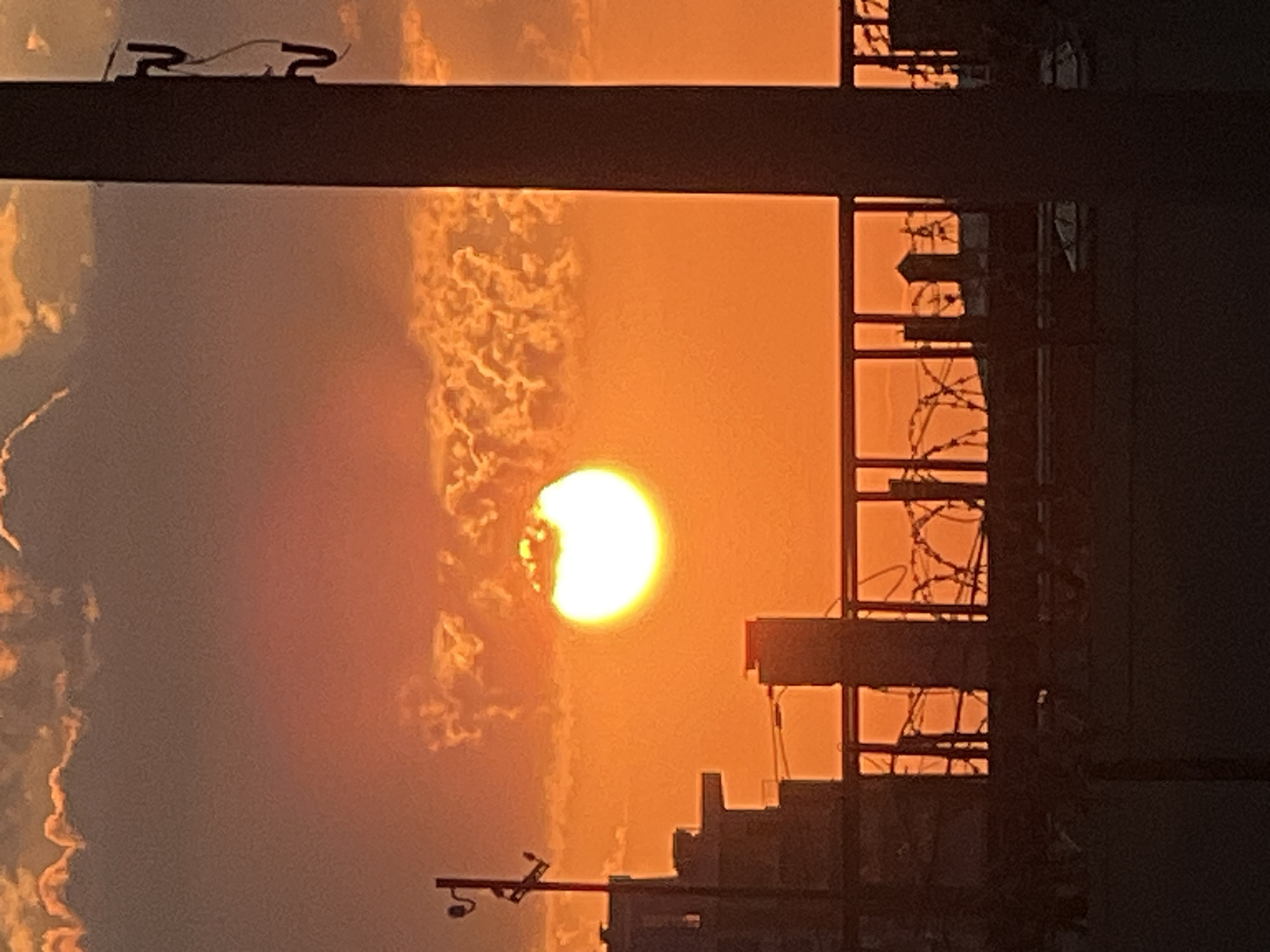 The image depicts a partial solar eclipse observed at sunset, likely in an urban setting given the presence of buildings and structures. The sun, partially obscured, casts a warm, orange glow across the sky, illuminating scattered clouds.
In the foreground, a dark, horizontal bar suggests a window frame or a balcony railing. To the left of the sun, some cloudy formations are visible, with their edges brightly lit by the setting sun. To the right, the silhouette of a building is prominent, with its windows appearing as dark rectangles. Within these windows, intricate patterns of barbed wire are discernible, suggesting a secured area or a barrier. Further to the right and in the background, more building structures are visible in silhouette, their details lost in the dim light.
At the very top of the image, on the dark horizontal bar, a small object with two prongs and a wire attached can be seen, possibly part of an antenna or a small fixture. At the bottom left, another silhouetted structure with a camera-like object is visible, also mounted on a horizontal bar.
The overall scene evokes a sense of an urban environment at dusk, with the unusual phenomenon of a solar eclipse adding a unique element. The warm hues of the sunset create a visually striking contrast with the dark, silhouetted structures. The presence of barbed wire hints at a possibly industrial or restricted zone. The time of day is clearly sunset, indicated by the color of the sky and the position of the sun. The weather appears clear enough to observe the eclipse, though clouds are present. The location context provided is Krasnodar, Russia. No visible text is present in the image.