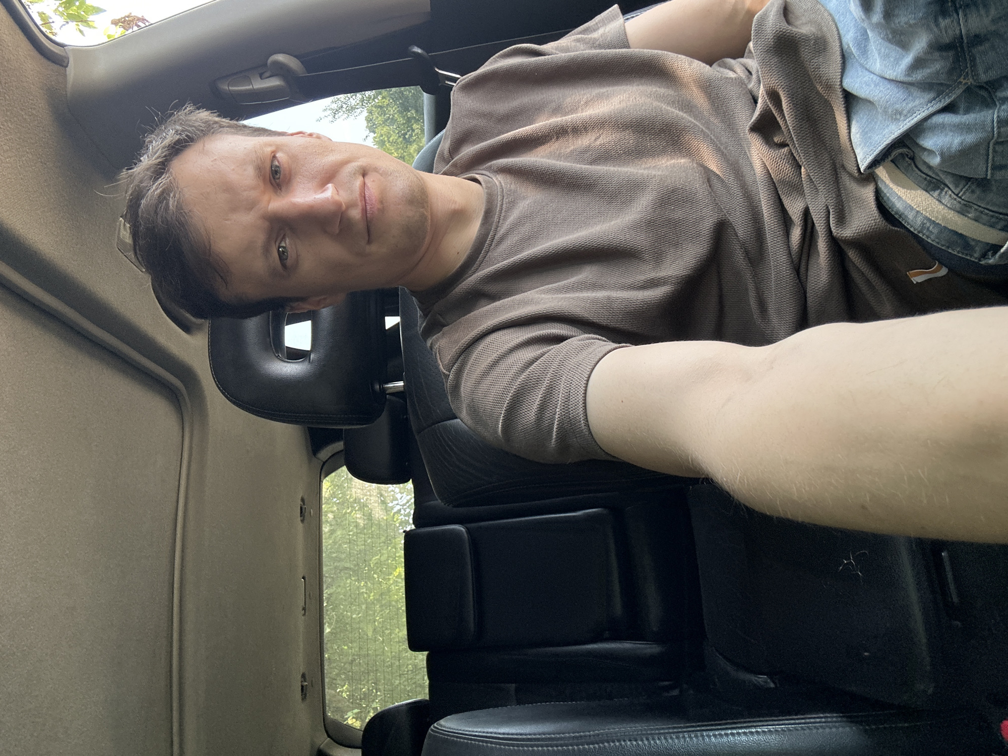 A man is seated in the back seat of a car. He is wearing a brown t-shirt and blue jeans. The car has black leather seats and a sunroof. The background is blurred trees, suggesting an outdoor setting, possibly in Makiv, Ukraine. The man is looking at the camera and has his arm extended, as if taking a selfie.