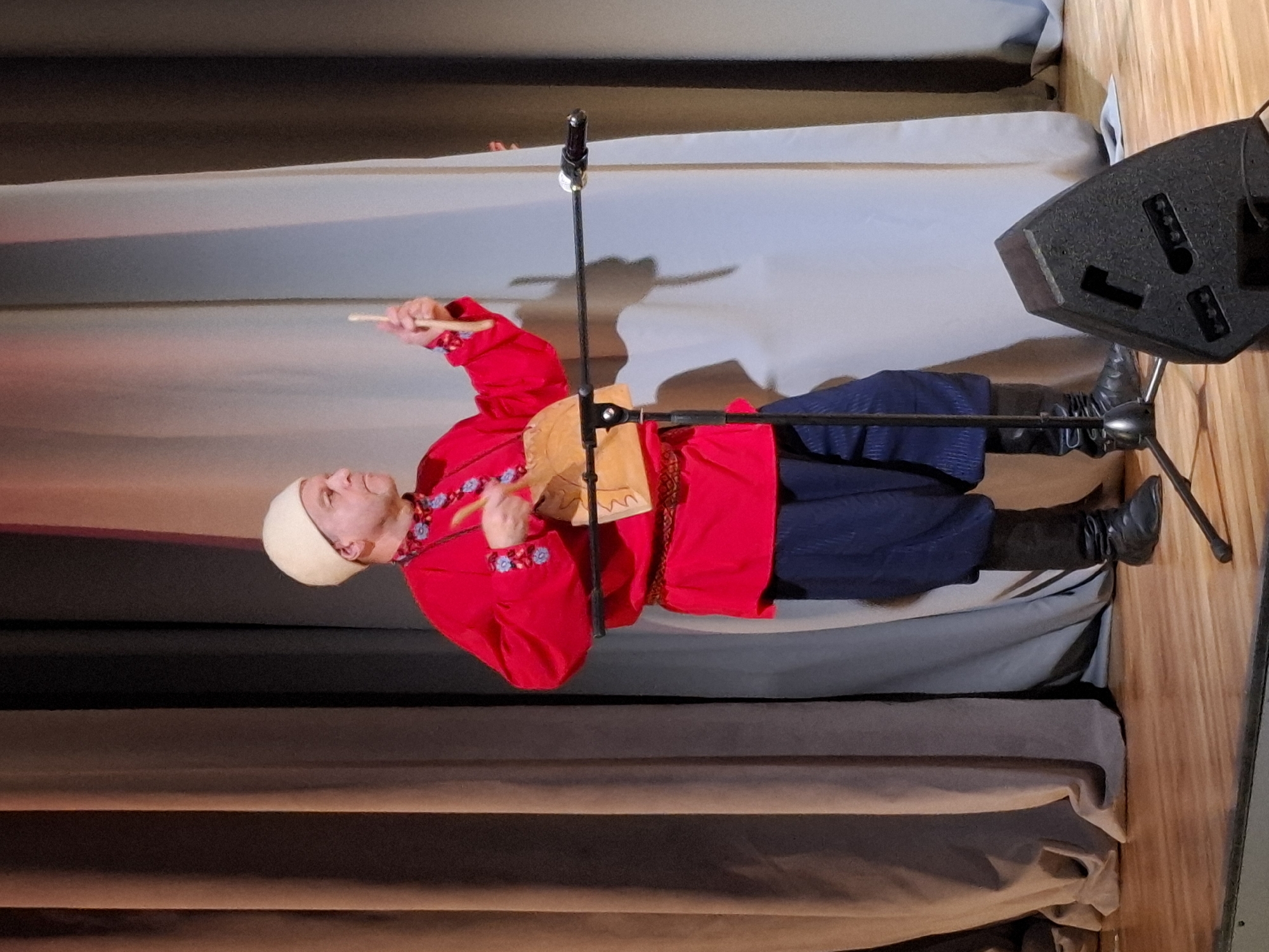 A man in traditional Russian attire stands on a wooden stage in front of a backdrop of curtains. He is wearing a red tunic with blue embroidered accents, a red belt, dark blue pants, black boots, and a white skullcap. He holds a wooden drum and a beater in his hands, appearing to be in the middle of a performance. A microphone stand is positioned in front of him, and a black speaker is visible in the foreground. The image was taken in Kungur, Russia.
