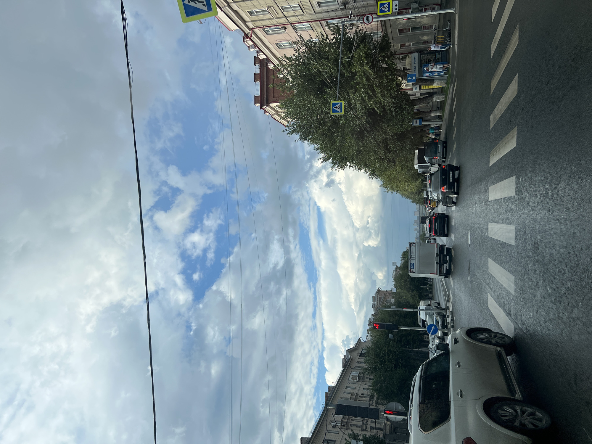The image depicts a street scene in Novosibirsk, Russia, under a cloudy sky. The perspective is from inside a vehicle, looking forward. The street is wet, suggesting recent rain, and features multiple lanes of traffic with cars and trucks. Several pedestrian crossings are visible, marked by white stripes.
On the left side of the street, there are apartment buildings and trees. A traffic light shows a red signal for pedestrians. On the right side, more apartment buildings line the street, with trees and shops visible at street level. Pedestrian crossing signs are posted.
Several people are present on the sidewalks, some walking and others standing. Electric scooters are parked on the sidewalk. The overall atmosphere is that of a typical urban environment on a possibly overcast day.