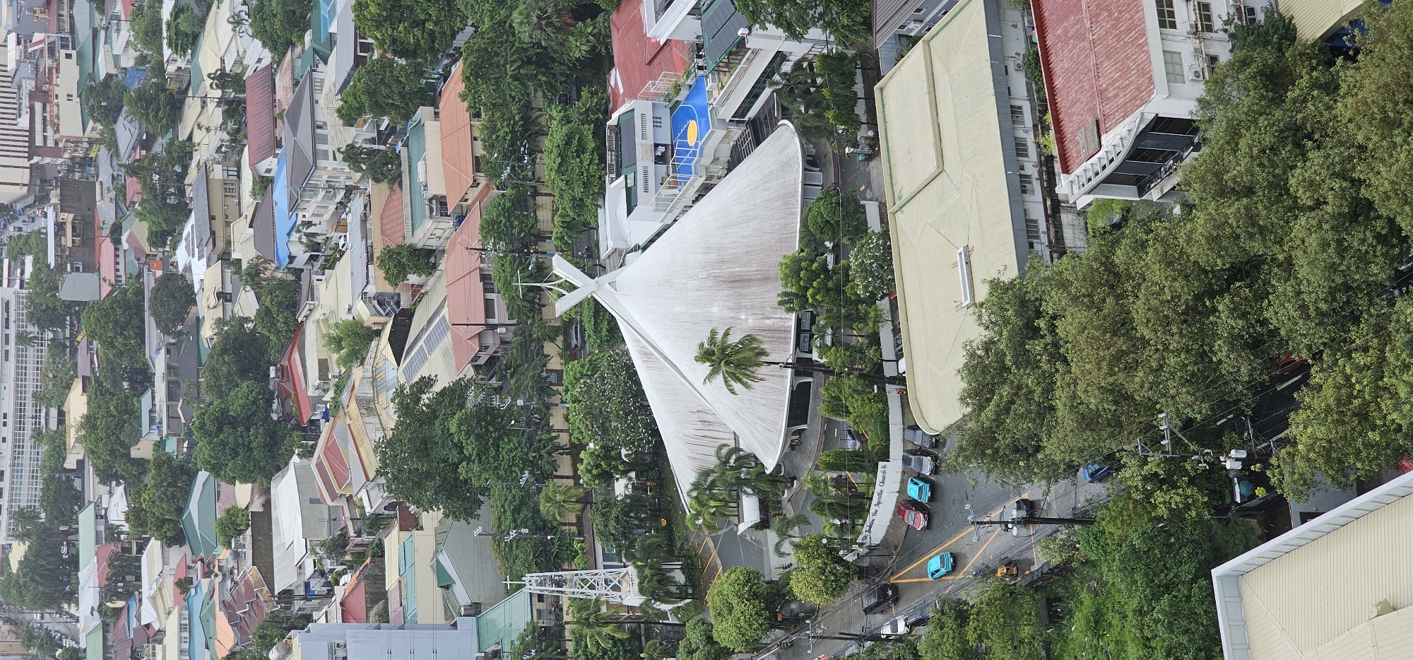 An aerial, eye-level shot captures a sprawling urban landscape in Makati, Philippines, under an overcast sky. The prominent feature is a large, white, tent-like structure, possibly a church or assembly hall, with a distinctive cross at its apex. Adjacent to this structure is a tall, white, lattice-like tower topped with a cross. The surrounding area is densely populated with a variety of buildings, including residential homes with colorful roofs and a larger, modern building with a blue and yellow court on its rooftop. A street with several parked cars, including two blue sedans and a black SUV, runs alongside the church. Lush green trees are interspersed throughout the scene, creating a contrast between the built environment and nature. Power lines and poles are visible, suggesting a functional urban infrastructure. The overall impression is one of a developed urban area with both religious and residential elements.