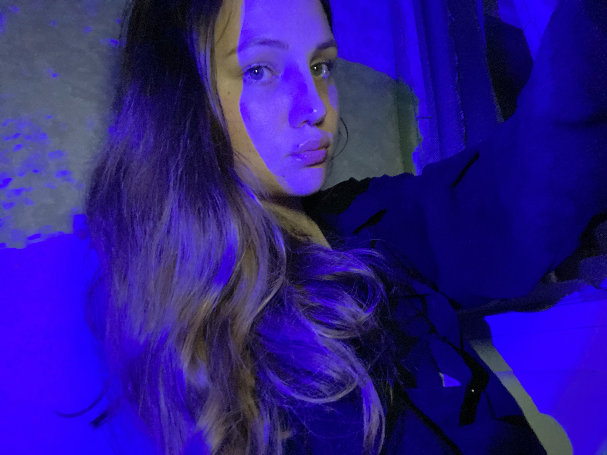 A young woman is pictured in a selfie, bathed in strong blue and yellow lighting. She has long, wavy hair with lighter ends. She is wearing a black jacket. The location is not discernible from the image, and therefore cannot be connected to Izhevsk, Russia.