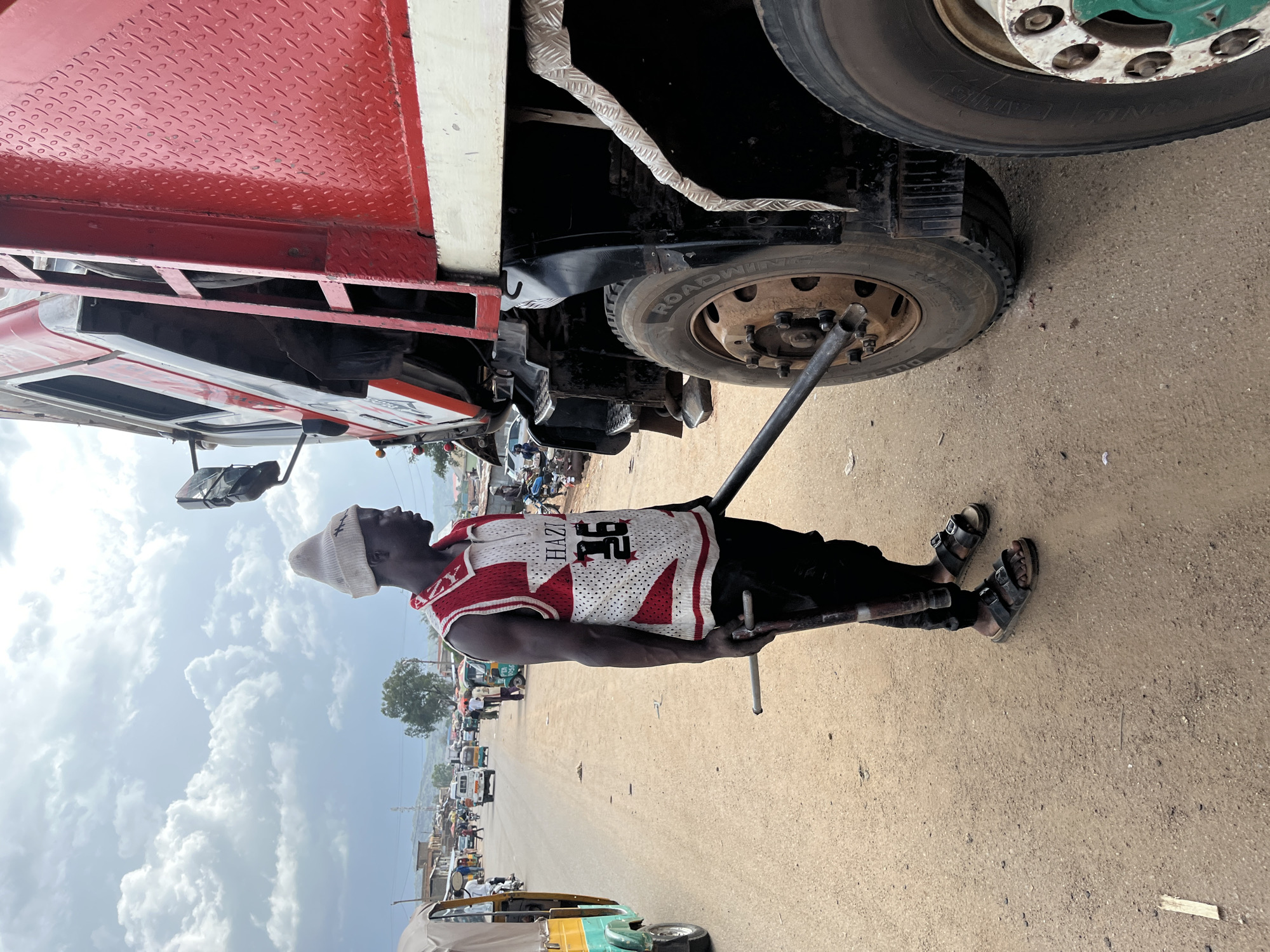 The image shows a man changing a tire on a large red truck. He is wearing a white beanie, a red and white basketball jersey with the number 36 and the name "HAZI" on it, black pants, and black sandals. He is using a jack to lift the truck and a lug wrench to loosen the bolts on the tire. The scene appears to be a roadside in Jos, Nigeria. In the background, there are other vehicles, including what appear to be minibuses and motorcycles, and buildings, suggesting a busy marketplace or town. The sky is partly cloudy, indicating it is daytime. The ground is a dusty, sandy surface. The overall impression is one of everyday life and work taking place in a community setting.