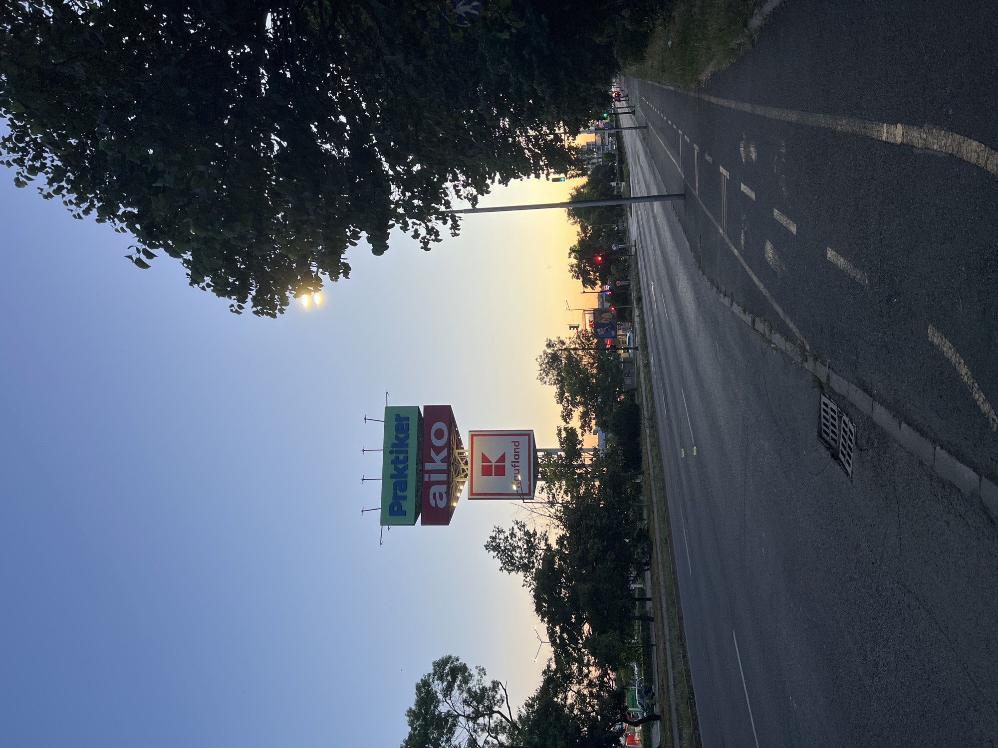 The image shows a street scene at dusk in Burgas, Bulgaria. A large billboard with three signs is visible, advertising "Praktiker," "aiko," and "Kaufland." The sky is a clear blue transitioning to orange at the horizon, indicating the setting sun. There is a paved road with traffic lanes and a separate bicycle lane marked with white lines. Trees line the side of the road, and streetlights are present. In the background, buildings and traffic signals can be seen. The overall impression is an urban environment at the end of the day.