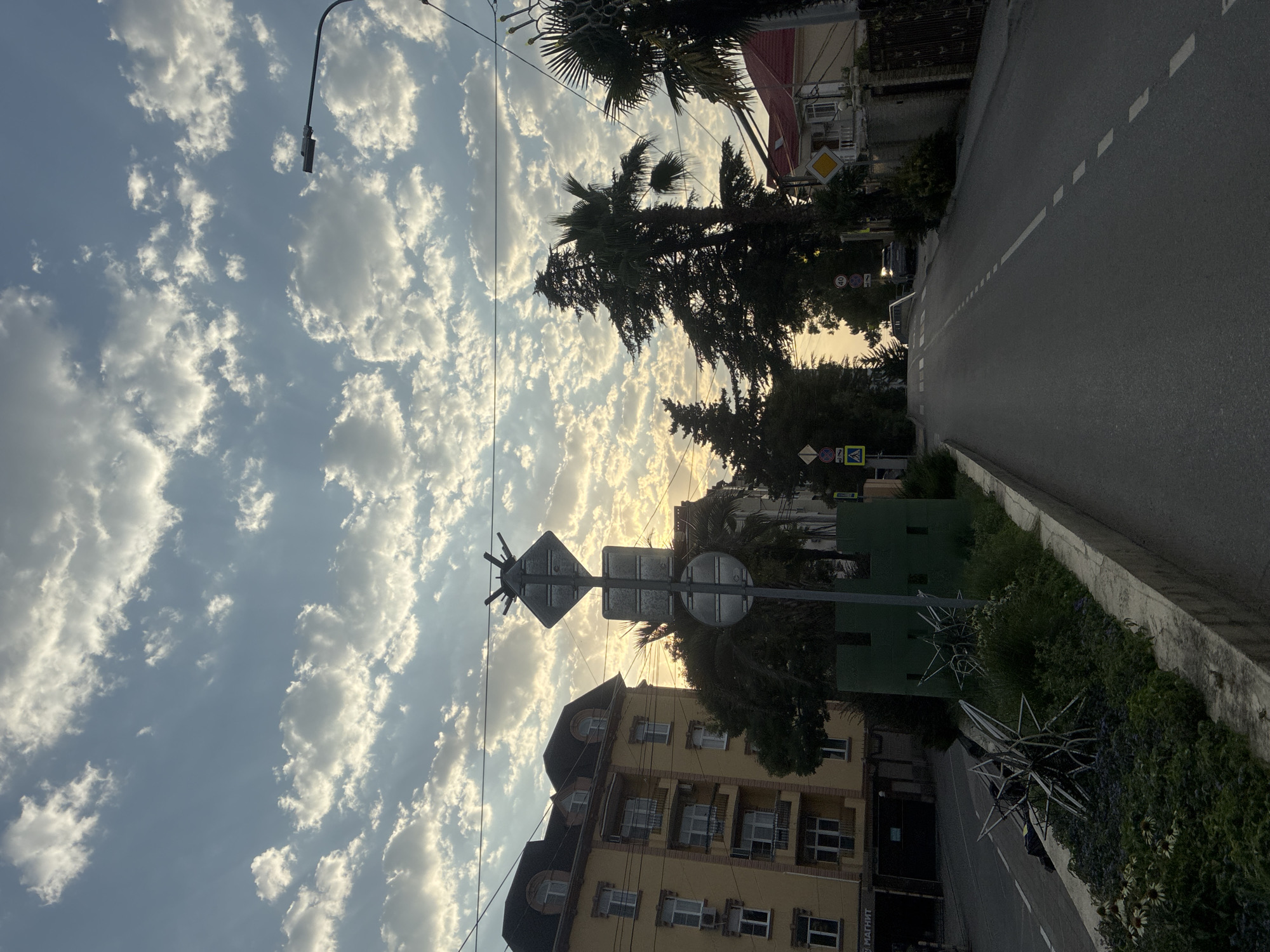 The image depicts a street scene in Sochi, Russia, during what appears to be late afternoon or early evening. The sky is filled with a dramatic display of cumulus clouds, illuminated by the setting sun, which creates a warm glow. A street lamp is visible on the right side of the frame, suggesting an urban or suburban setting.
On the left, a multi-story yellow building with a dark, mansard roof and balconies is prominent. Several windows are visible, some with air conditioning units. The building is set back from the street, with a sidewalk and a green, decorative border separating it from the road. Within this border, there are various plants, including flowers and what appear to be decorative metal sculptures resembling spiked spheres. A small, green, castle-like structure is also situated in the border.
The road itself is asphalt, with a white dashed line marking the center. There is a slight incline to the road, leading away from the viewer. On the right side of the road, a fence and a red-roofed house are visible. Trees, including palm trees and cypress trees, are scattered throughout the scene, adding to the Mediterranean feel often associated with Sochi.
Several traffic signs are present. One large signpost with multiple circular and diamond-shaped signs stands in the middle ground. Further down the road, additional signs indicating speed limits and pedestrian crossings are visible. Two cars are parked on the side of the road in the distance. The overall impression is a quiet, somewhat residential street bathed in the soft light of sunset, with distinctive local flora and architecture.