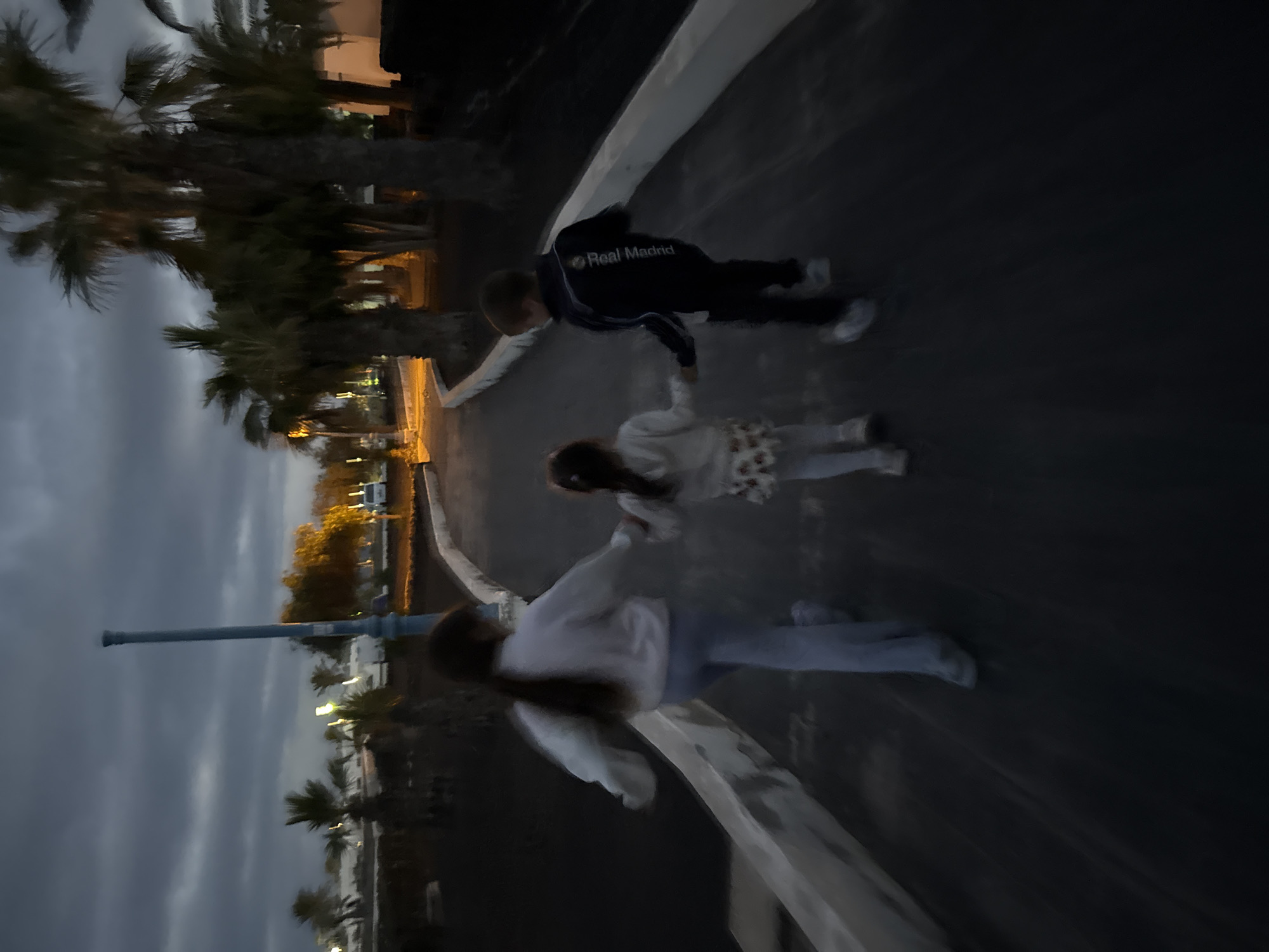 Three children walk hand-in-hand on a paved surface next to a white ledge. The first child, wearing a dark-colored tracksuit with "Real Madrid" printed on the back, holds hands with a second child, who in turn holds hands with a third child wearing a white hooded sweatshirt and light blue jeans. Palm trees line the left side of the image. Streetlights are visible in the background. The location is Tías, Spain.